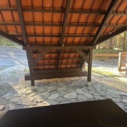 ADIKRISBAU Remonty i Wykończenia Krzysztof Smaza - Zadaszona wiata z drewnianą konstrukcją i pomarańczową dachówką, widok od wewnątrz. Na zewnątrz widoczny fragment żółtego samochodu i leśny krajobraz.