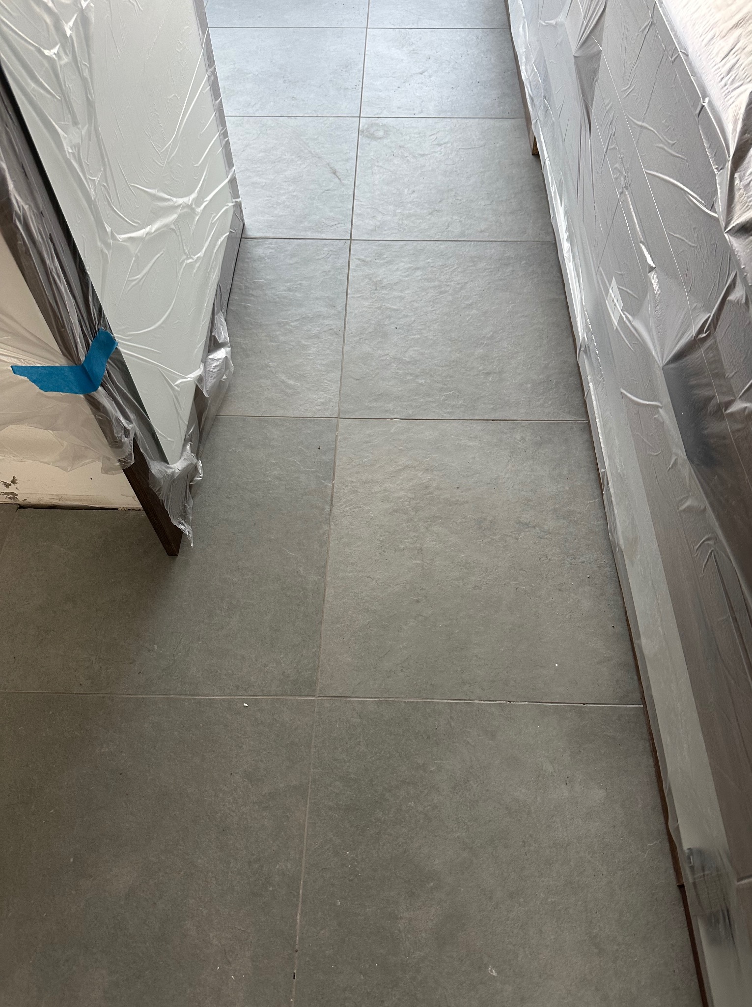 Szare płytki podłogowe z fugami, częściowo zakryte folią ochronną, prawdopodobnie zabezpieczającą nowe meble lub drzwi przed zabrudzeniem podczas prac wykończeniowych.