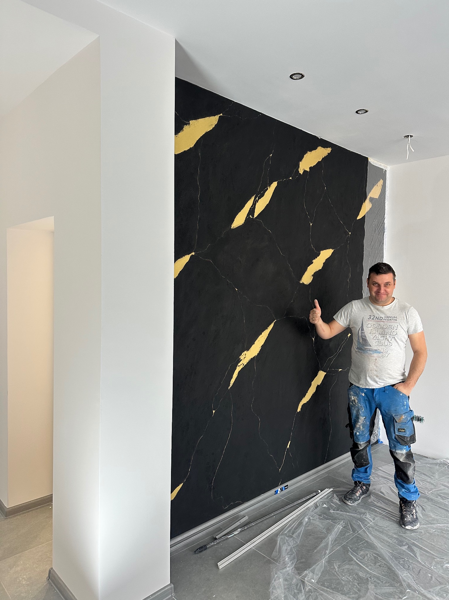Ściana z efektem czarnego marmuru i złotych żył, wykonana przez uśmiechniętego fachowca w roboczym stroju, z uniesionym kciukiem w górę. Podłoga zabezpieczona folią.