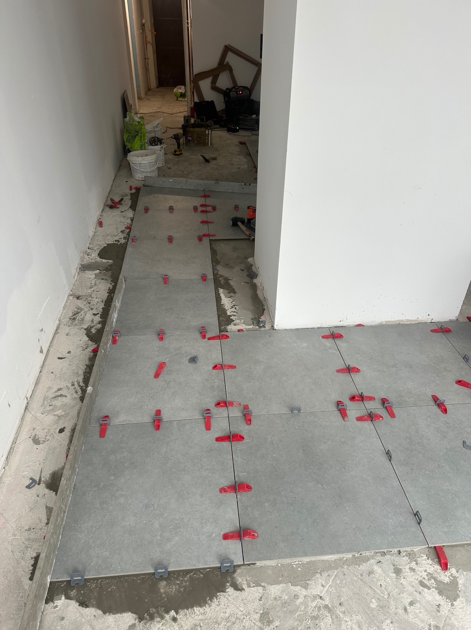 Układanie szarych płytek podłogowych z czerwonymi i szarymi klipsami poziomującymi w trakcie prac wykończeniowych. Widoczne narzędzia i materiały budowlane w tle.