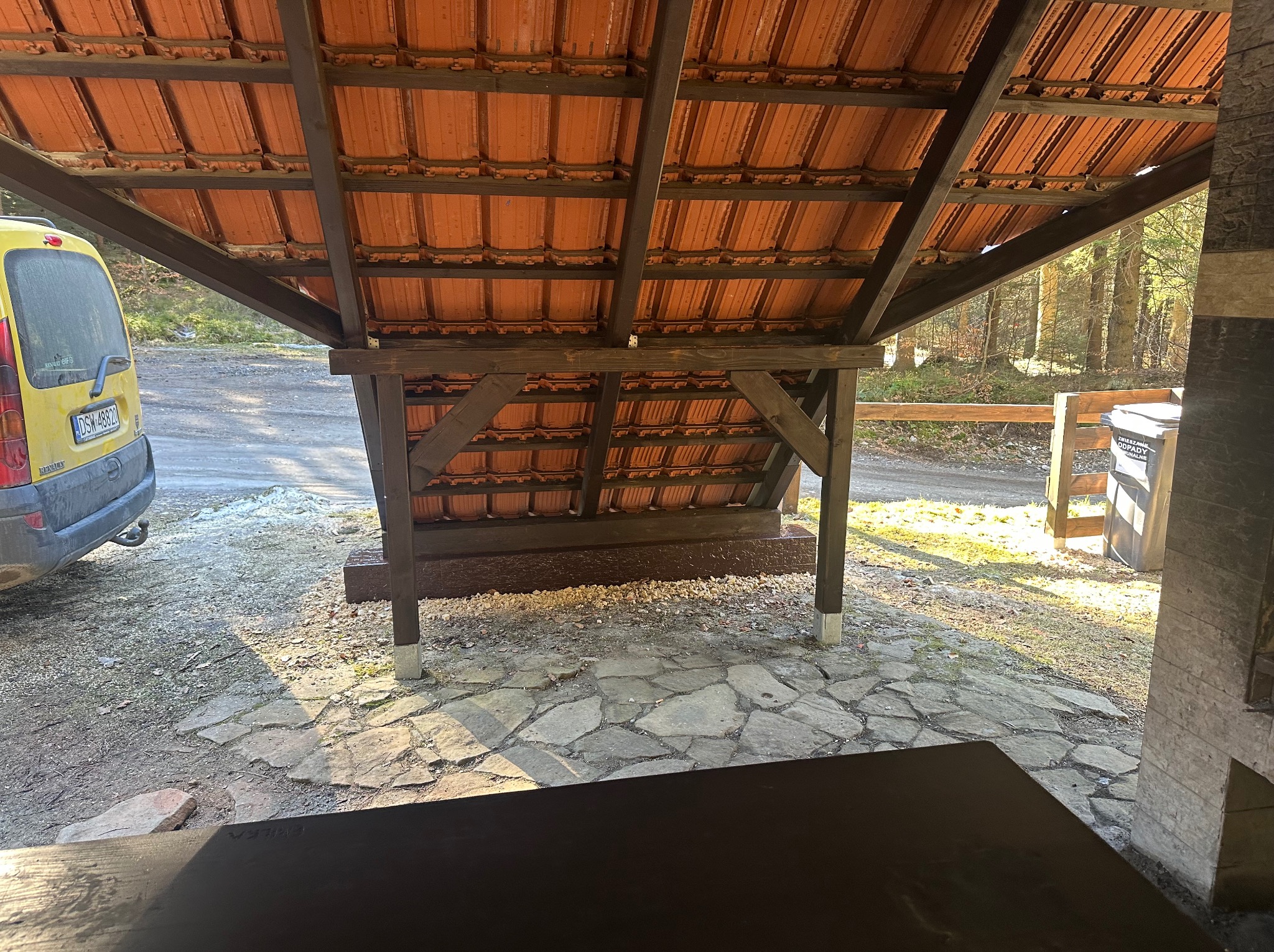 Zadaszona wiata z drewnianą konstrukcją i pomarańczową dachówką, widok od wewnątrz. Na zewnątrz widoczny fragment żółtego samochodu i leśny krajobraz.