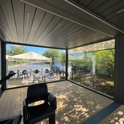 Ściany ze szkła - Zadaszony taras z drewnianą podłogą i szklanymi ścianami, widok na ogródek kawiarniany z parasolem i stolikami. Cień drzew na podłodze.