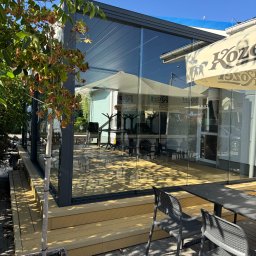 Ściany ze szkła - Szklana zabudowa tarasu z metalową ramą, widok z zewnątrz, z drewnianym podestem i stolikiem z krzesłami. W tle parasol reklamowy.