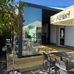 Ściany ze szkła - Szklana zabudowa tarasu z metalową ramą, widok z zewnątrz. Na pierwszym planie stolik i krzesła, obok drzewko w donicy. W tle widoczny parasol z logo.