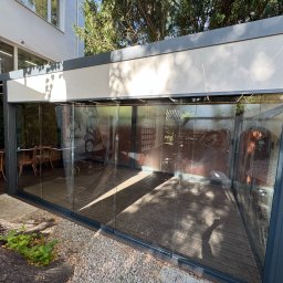 Ściany ze szkła - Nowoczesna, szklana zabudowa balkonu z metalową ramą, oświetleniem LED i drewnianą podłogą. Wnętrze z dekoracjami i meblami. Widok z zewnątrz, częściowo zasłonięty pniem drzewa.