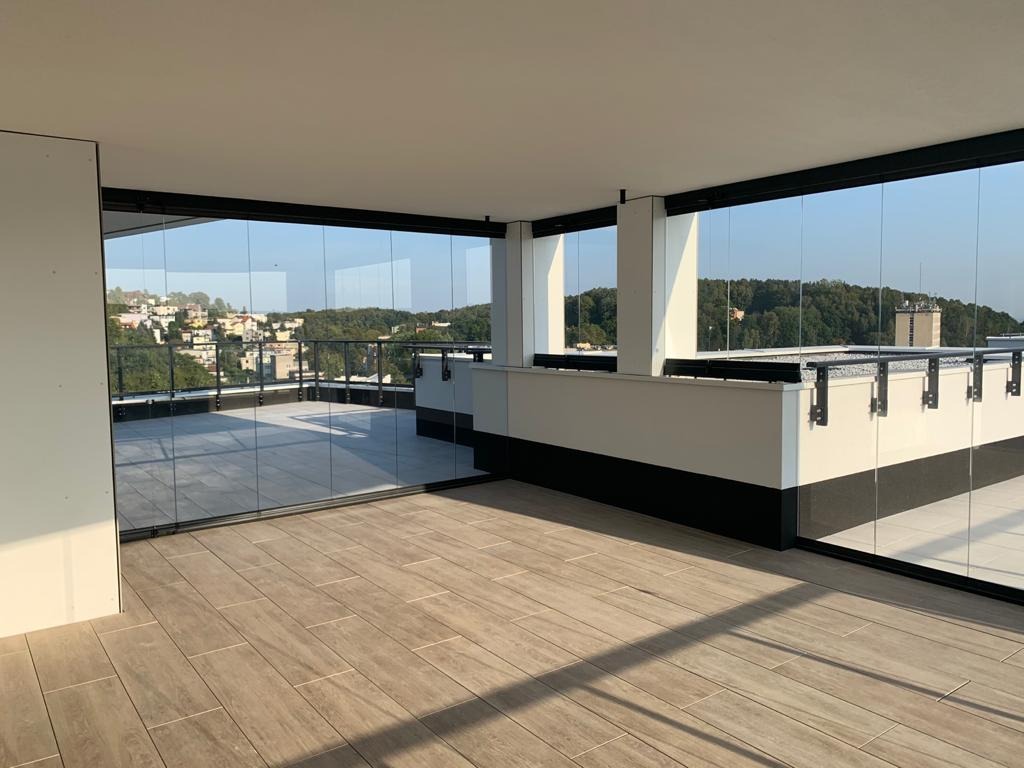Nowoczesna zabudowa balkonu z przesuwnymi szklanymi panelami, widok na miasto w tle, drewnopodobna podłoga, minimalistyczny design.