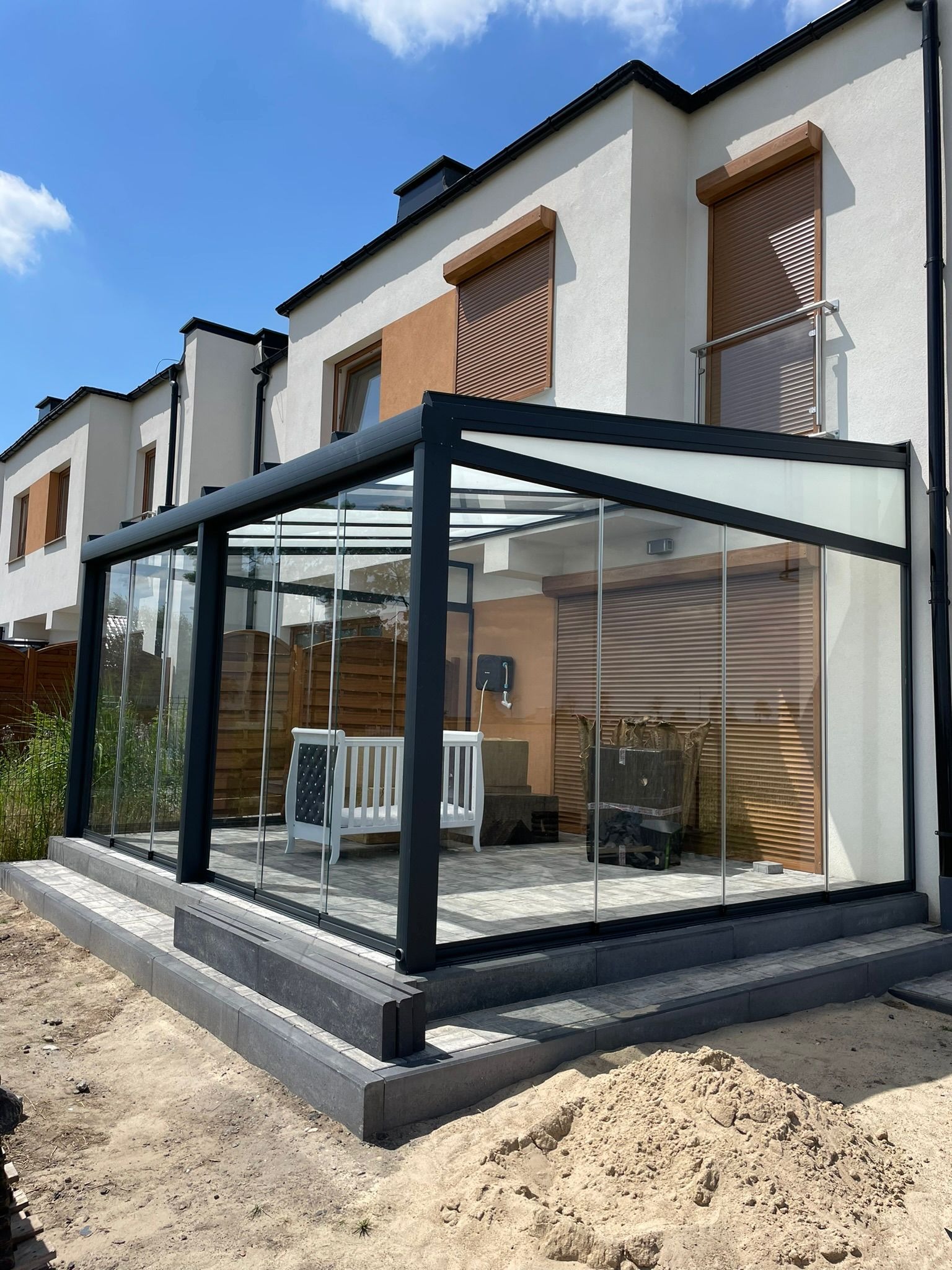 Nowoczesna, przeszklona konstrukcja z czarną ramą, przylegająca do budynku z roletami zewnętrznymi. Widoczne meble ogrodowe i elementy wykończeniowe.