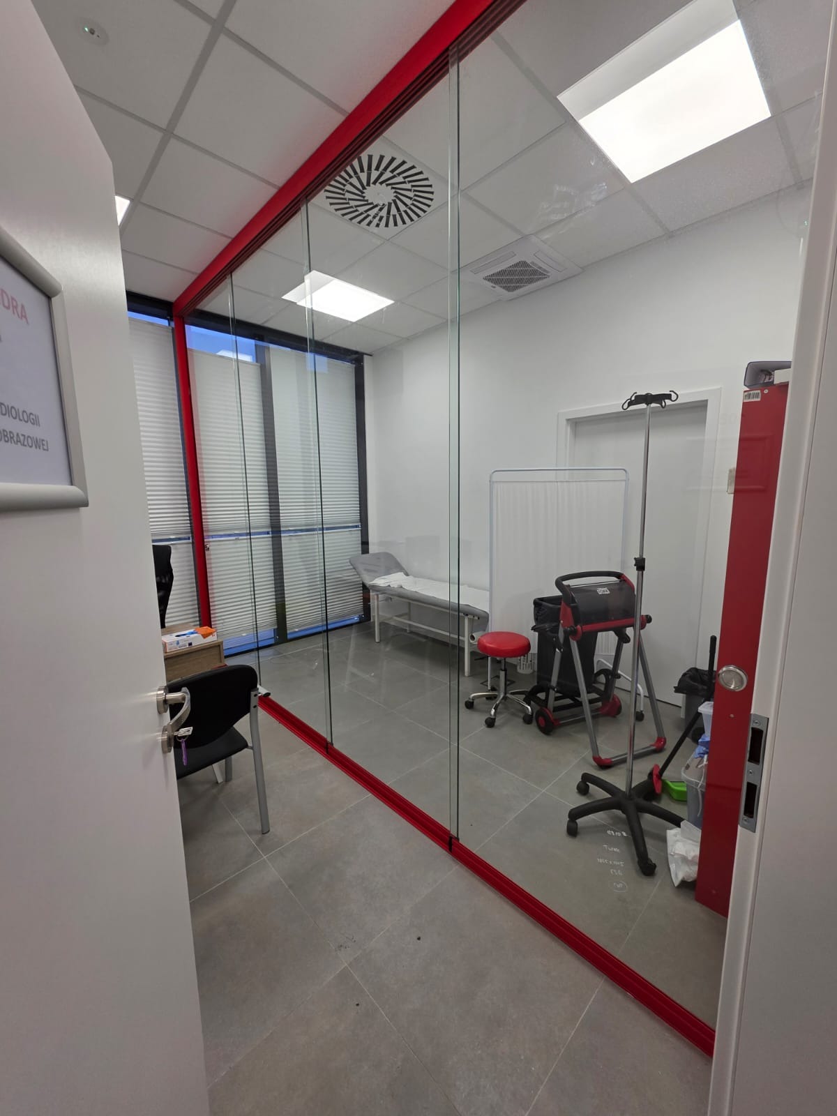 Szklana zabudowa z czerwonymi ramami oddzielająca gabinet lekarski z kozetką i sprzętem medycznym, widok z korytarza.