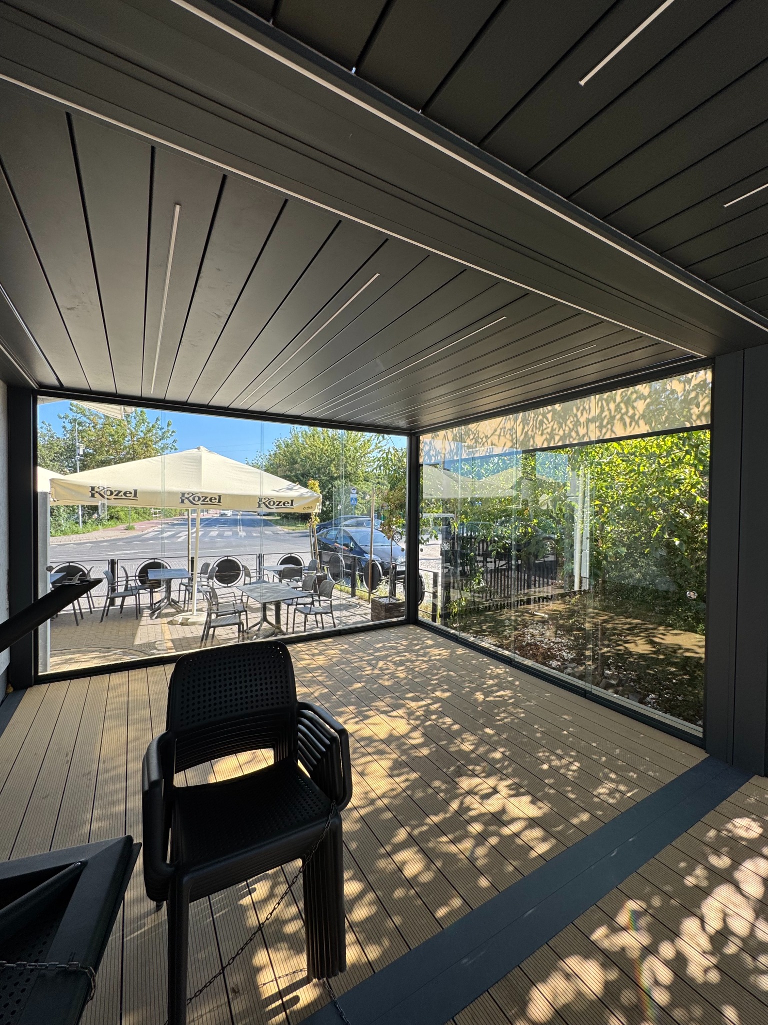 Zadaszony taras z drewnianą podłogą i szklanymi ścianami, widok na ogródek kawiarniany z parasolem i stolikami. Cień drzew na podłodze.