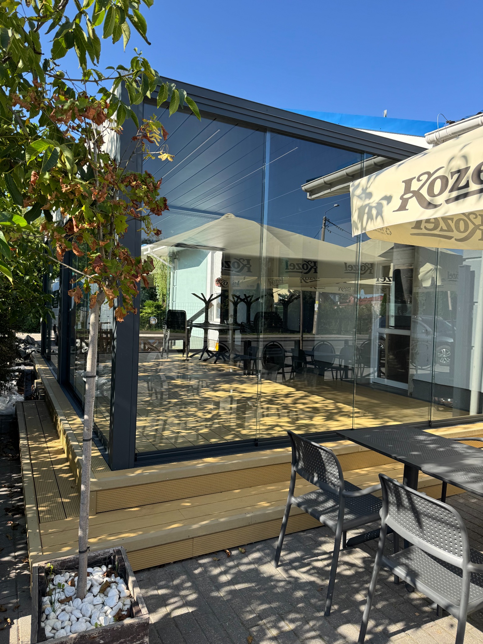 Szklana zabudowa tarasu z metalową ramą, widok z zewnątrz, z drewnianym podestem i stolikiem z krzesłami. W tle parasol reklamowy.