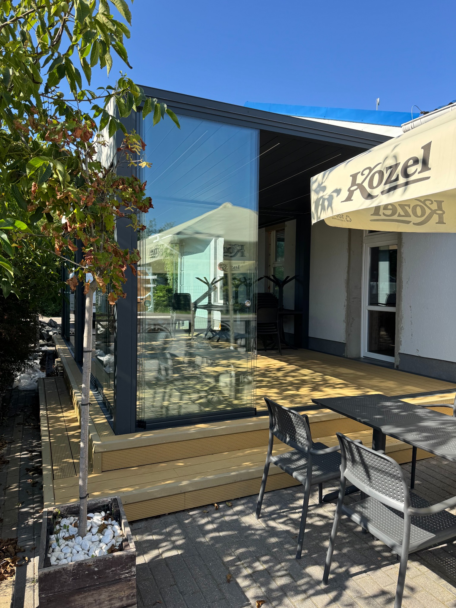 Szklana zabudowa tarasu z metalową ramą, widok z zewnątrz. Na pierwszym planie stolik i krzesła, obok drzewko w donicy. W tle widoczny parasol z logo.