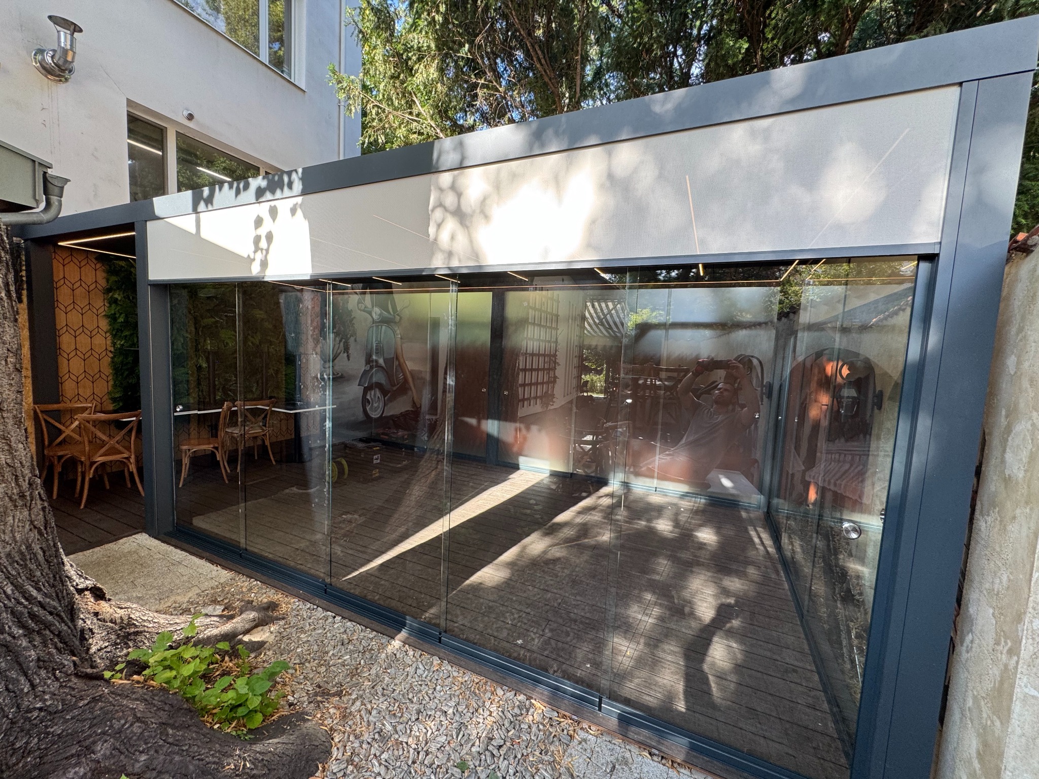 Nowoczesna, szklana zabudowa balkonu z metalową ramą, oświetleniem LED i drewnianą podłogą. Wnętrze z dekoracjami i meblami. Widok z zewnątrz, częściowo zasłonięty pniem drzewa.