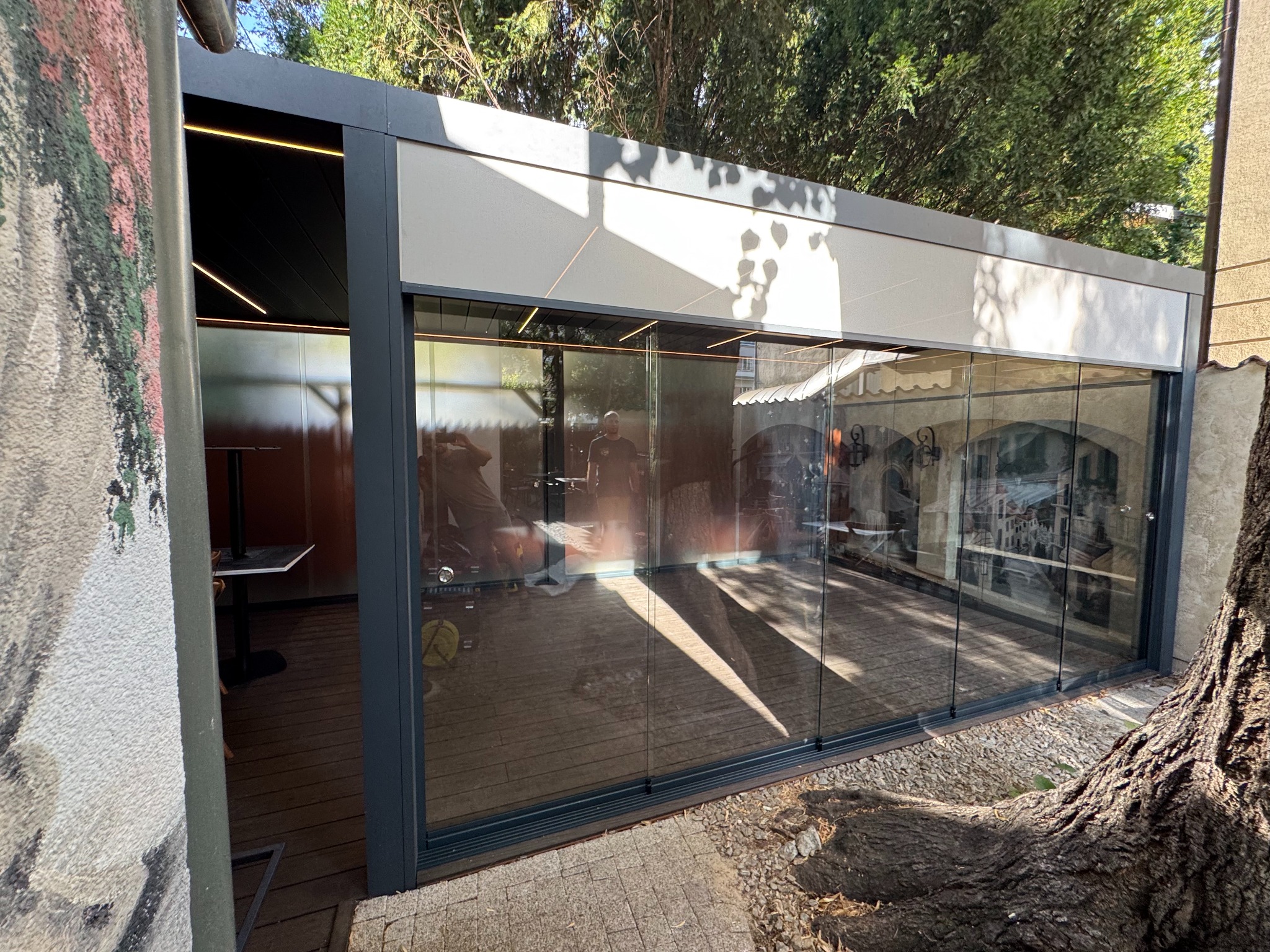 Szklana zabudowa tarasu z metalową ramą, widok z zewnątrz, z fragmentem pnia drzewa w prawym dolnym rogu i ścianą z muralem po lewej stronie.