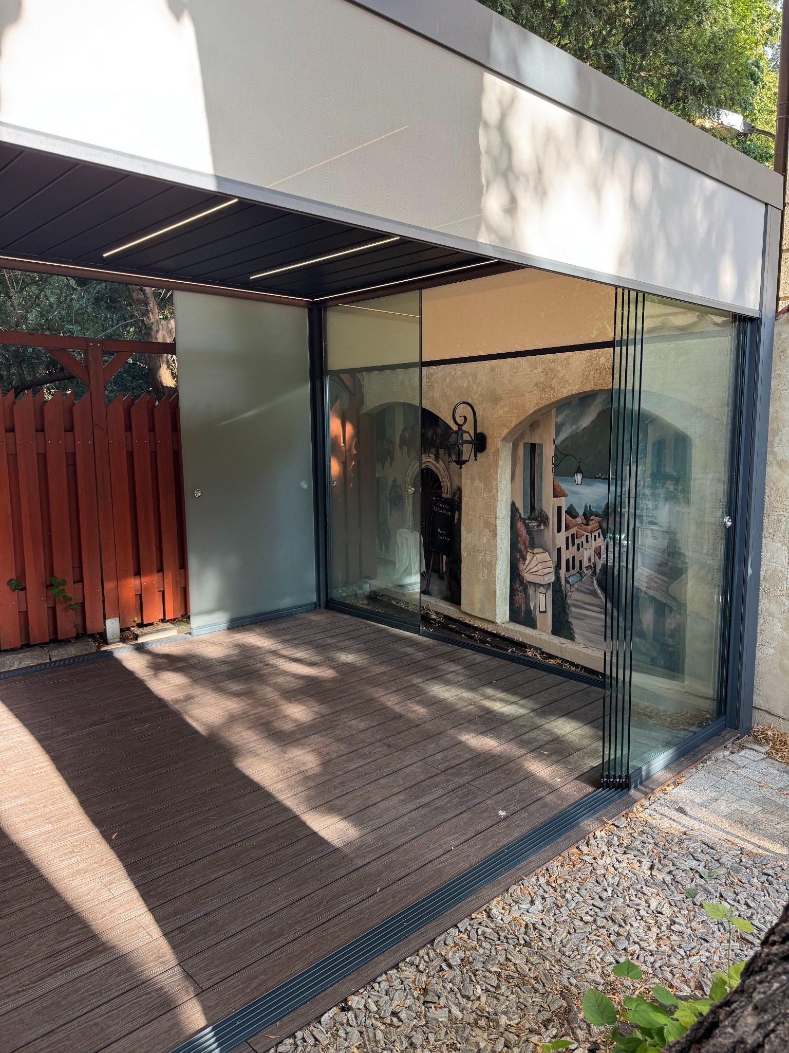 Zadaszona konstrukcja z przesuwnymi szklanymi panelami, drewnianą podłogą i dekoracyjną ścianą z motywem włoskiego miasteczka.