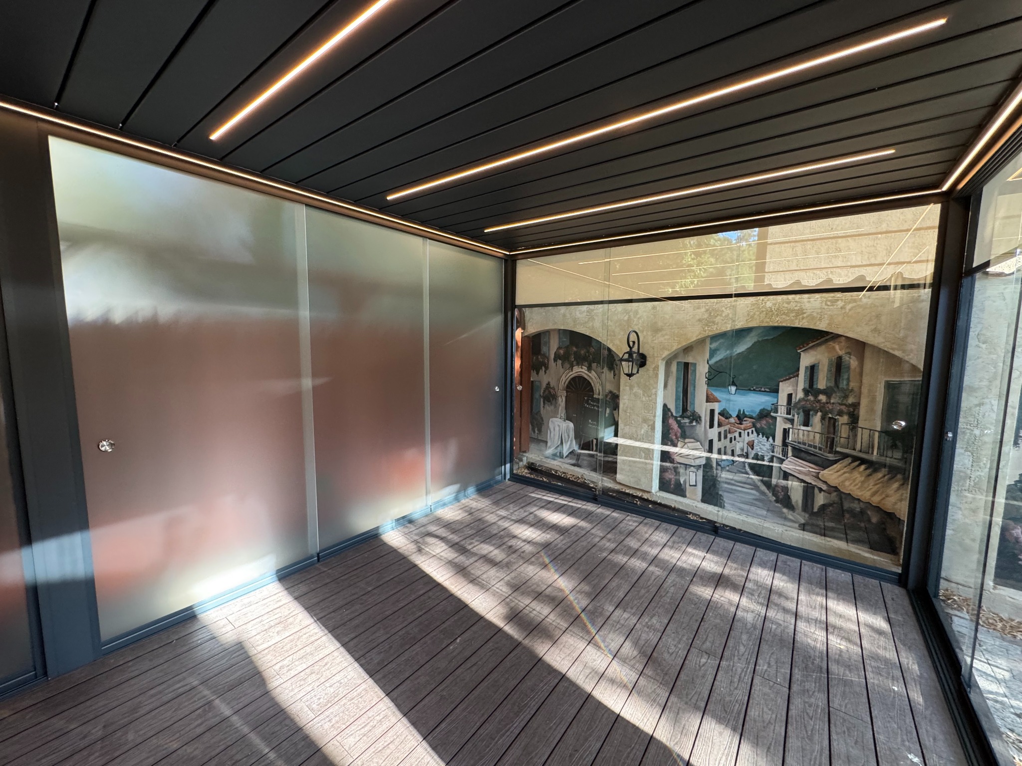 Nowoczesna zabudowa balkonu z przesuwnymi panelami z matowego szkła i drewnianą podłogą, oświetlona ledowymi listwami, z widokiem na mural z włoskim miasteczkiem.