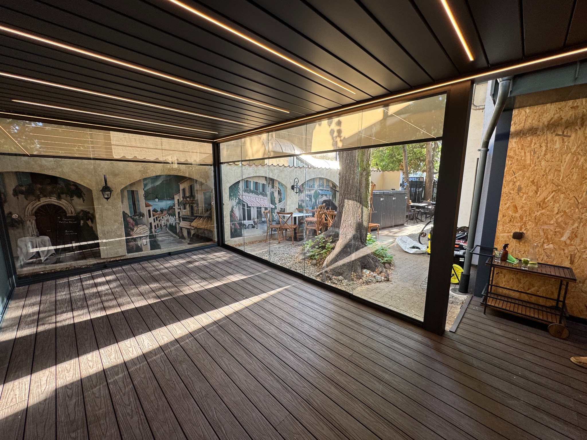 Zadaszony taras z drewnianą podłogą i szklanymi ścianami z malowidłami, oświetlony ledami, z widokiem na drzewo i ogród.