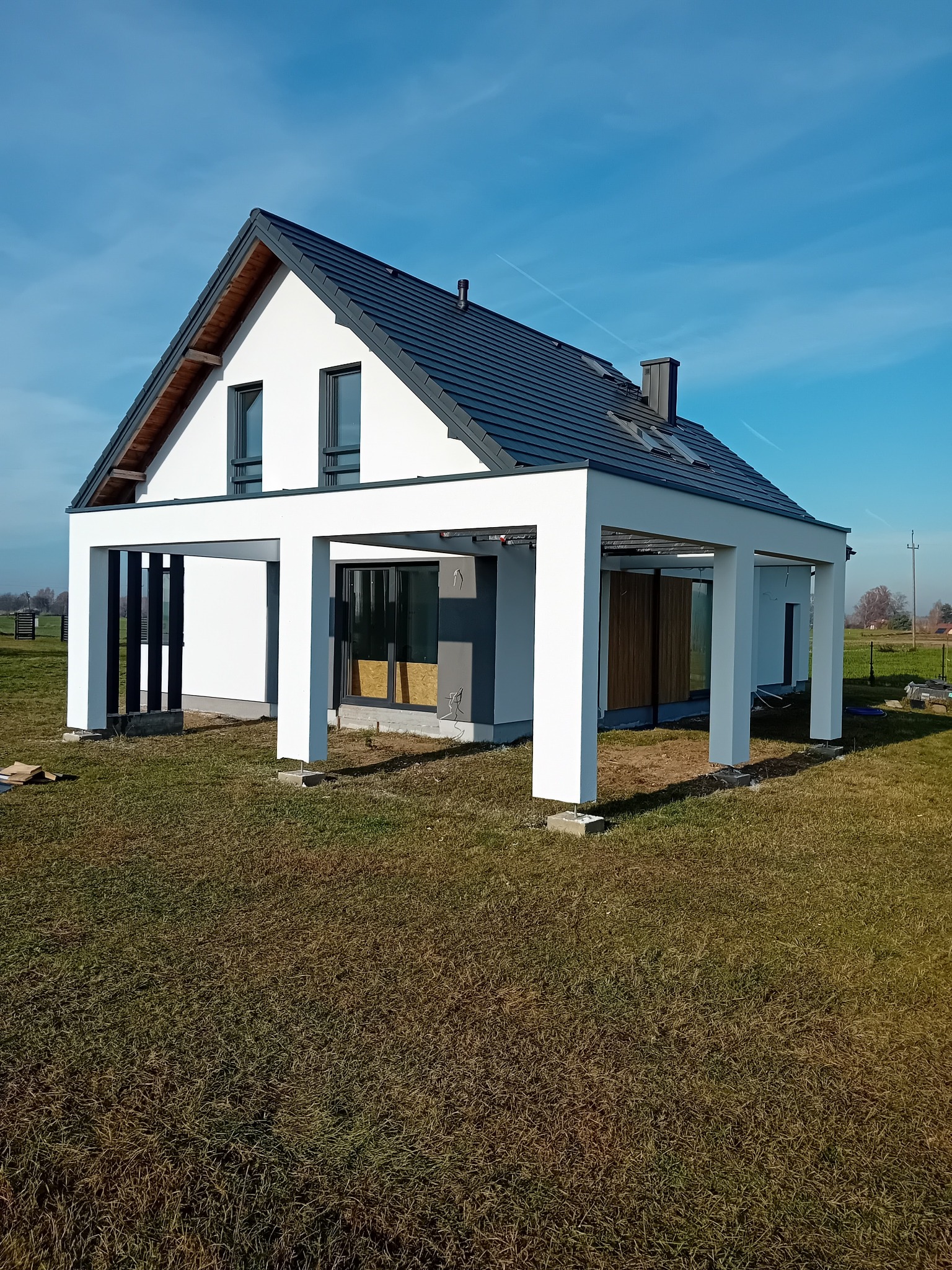 Nowoczesny, parterowy dom z płaskim dachem i białymi ścianami, otoczony zielonym trawnikiem pod błękitnym niebem. Widoczne elementy wykończeniowe i minimalistyczny design.