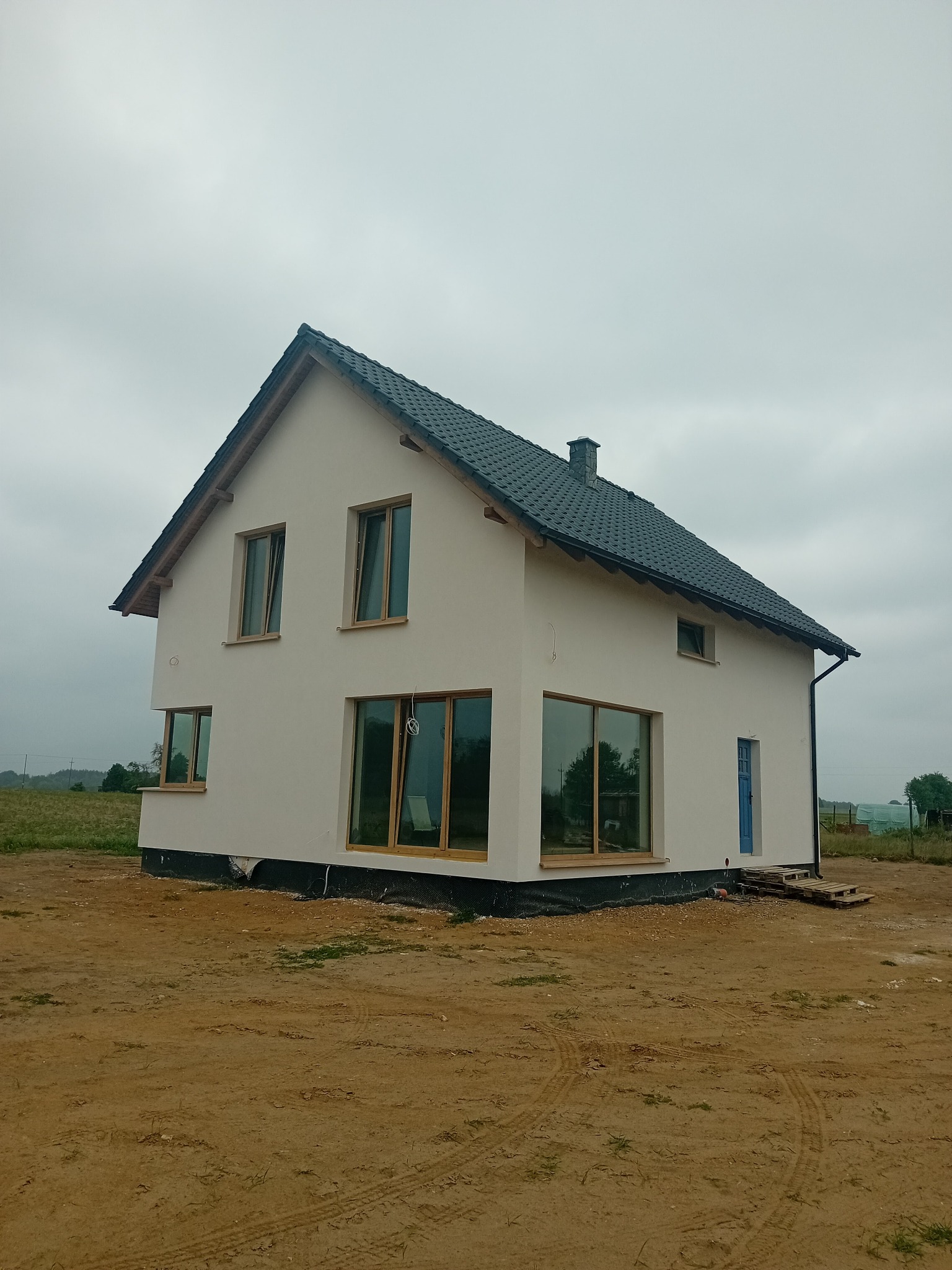 Nowoczesny dom jednorodzinny z jasną elewacją, ciemnym dachem i dużymi oknami, w trakcie wykończenia, na tle pochmurnego nieba i polnego krajobrazu.