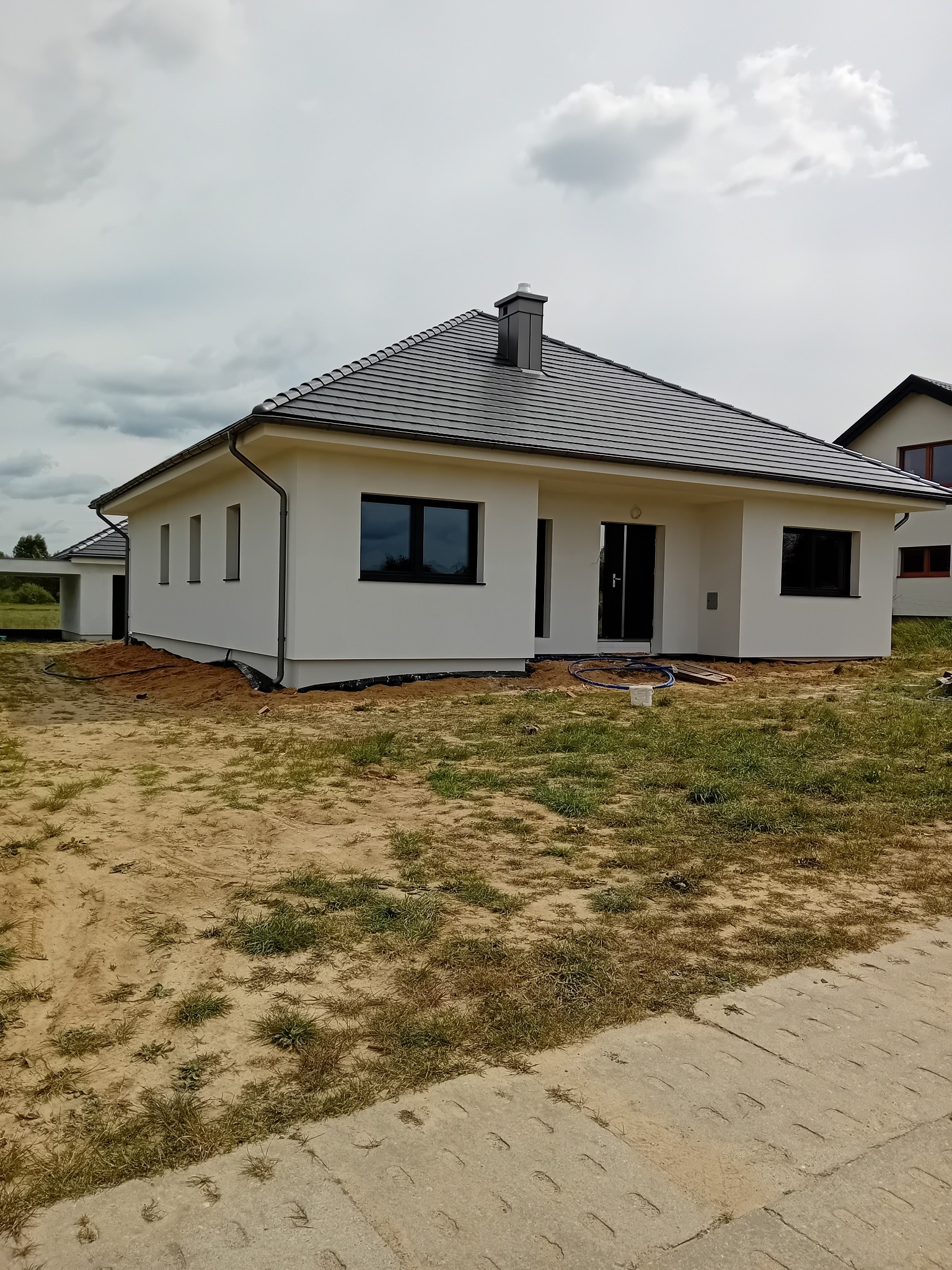 Nowoczesny, parterowy dom z ciemną dachówką i białymi ścianami, widoczny z zewnątrz, z elementami wykończeniowymi w trakcie realizacji. Uwagę zwraca minimalistyczny styl.