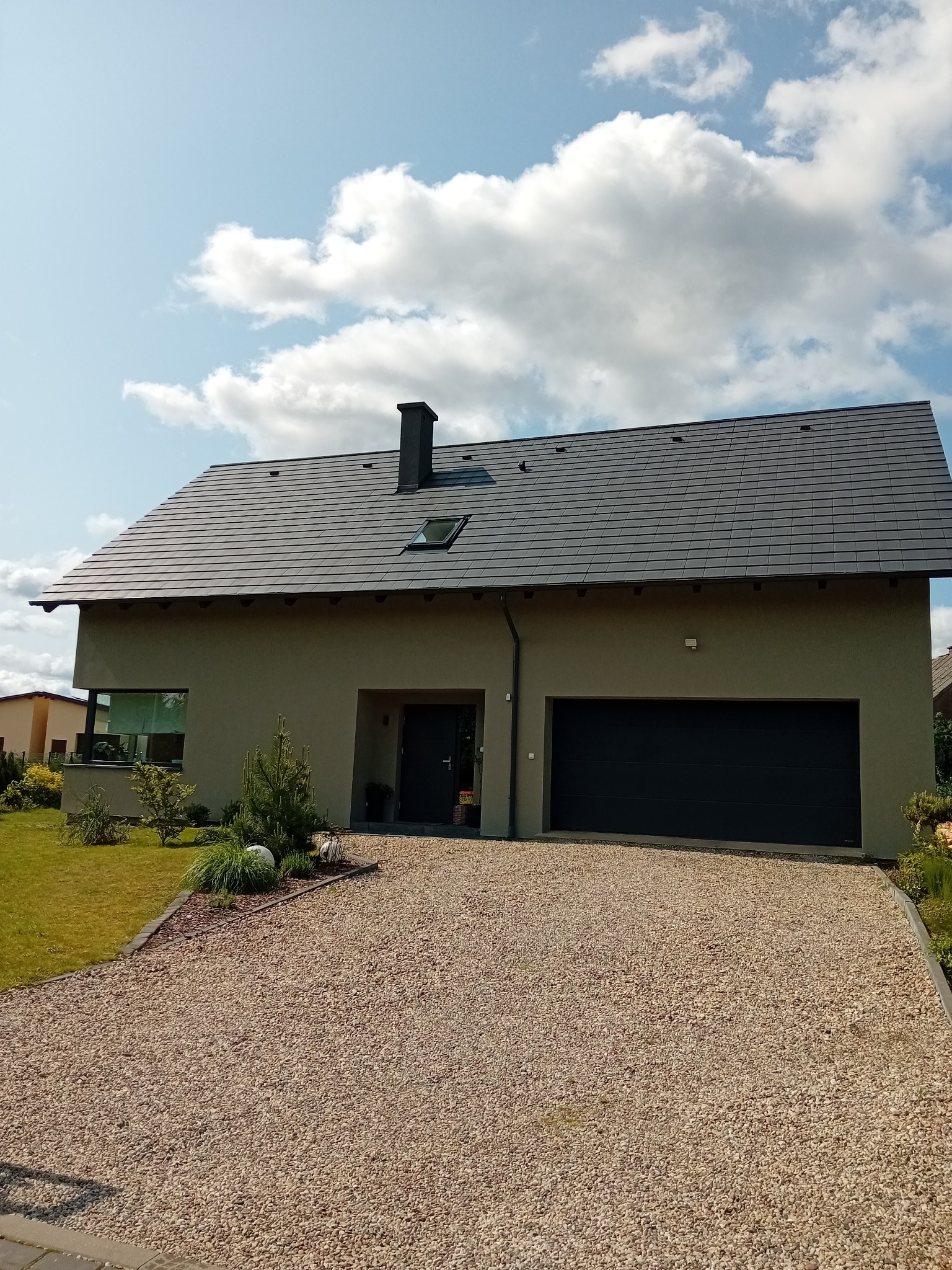 Nowoczesny dom jednorodzinny z grafitową dachówką i garażem, otoczony zielonym trawnikiem i żwirowym podjazdem w słoneczny dzień.