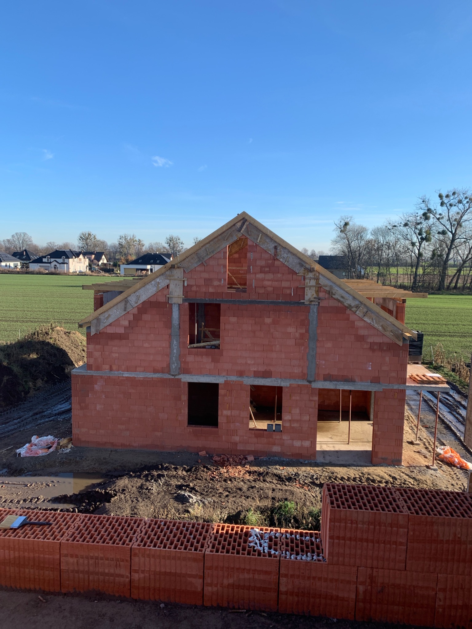 Wznoszenie domu z czerwonej cegły ceramicznej na tle zielonego pola i błękitnego nieba. Widoczne otwory okienne i drzwiowe, oraz elementy konstrukcji dachu.