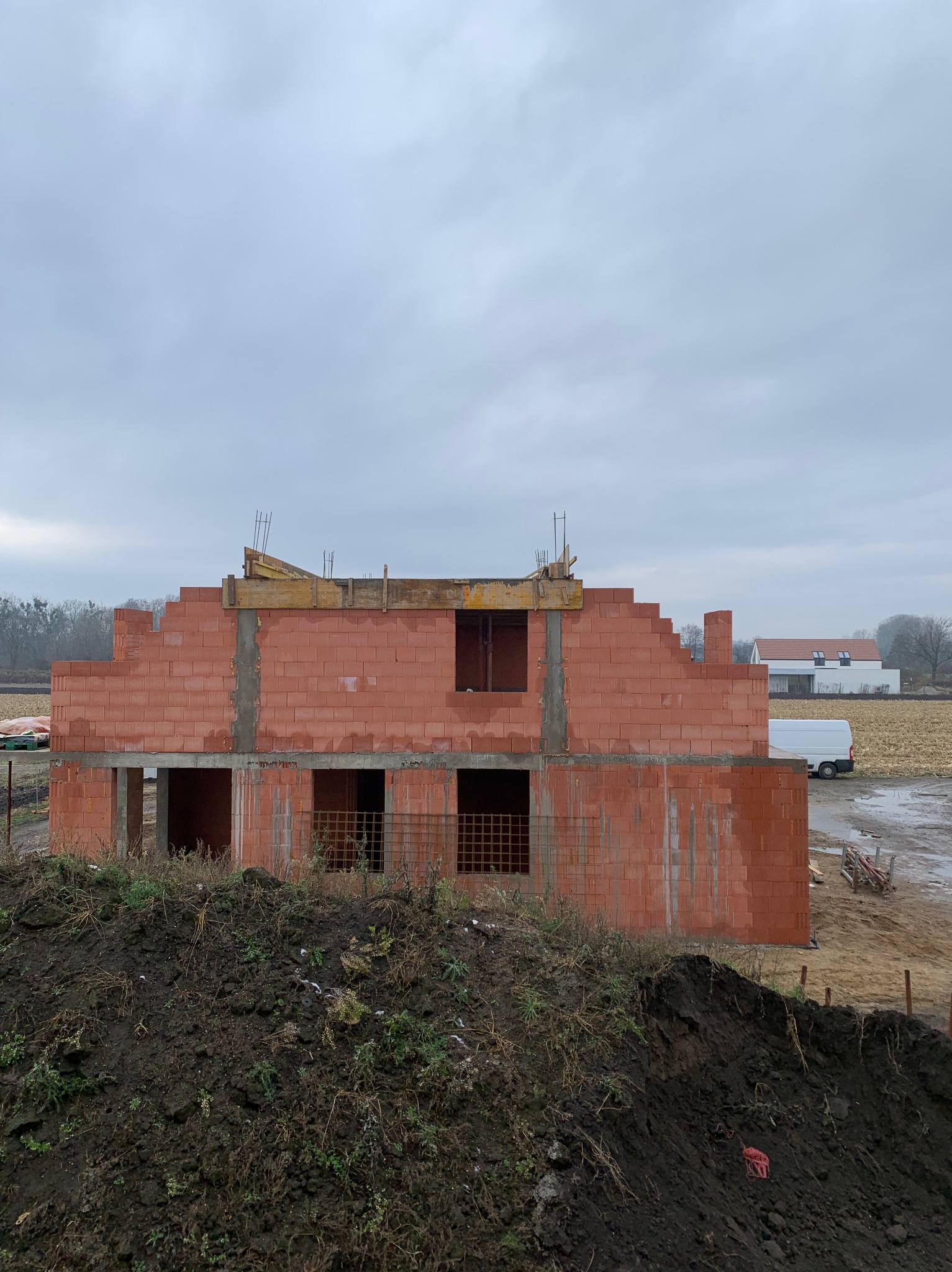 Surowy stan dwukondygnacyjnego domu z czerwonej cegły, z widocznymi otworami okiennymi i betonowymi słupami, na tle pochmurnego nieba i pola.