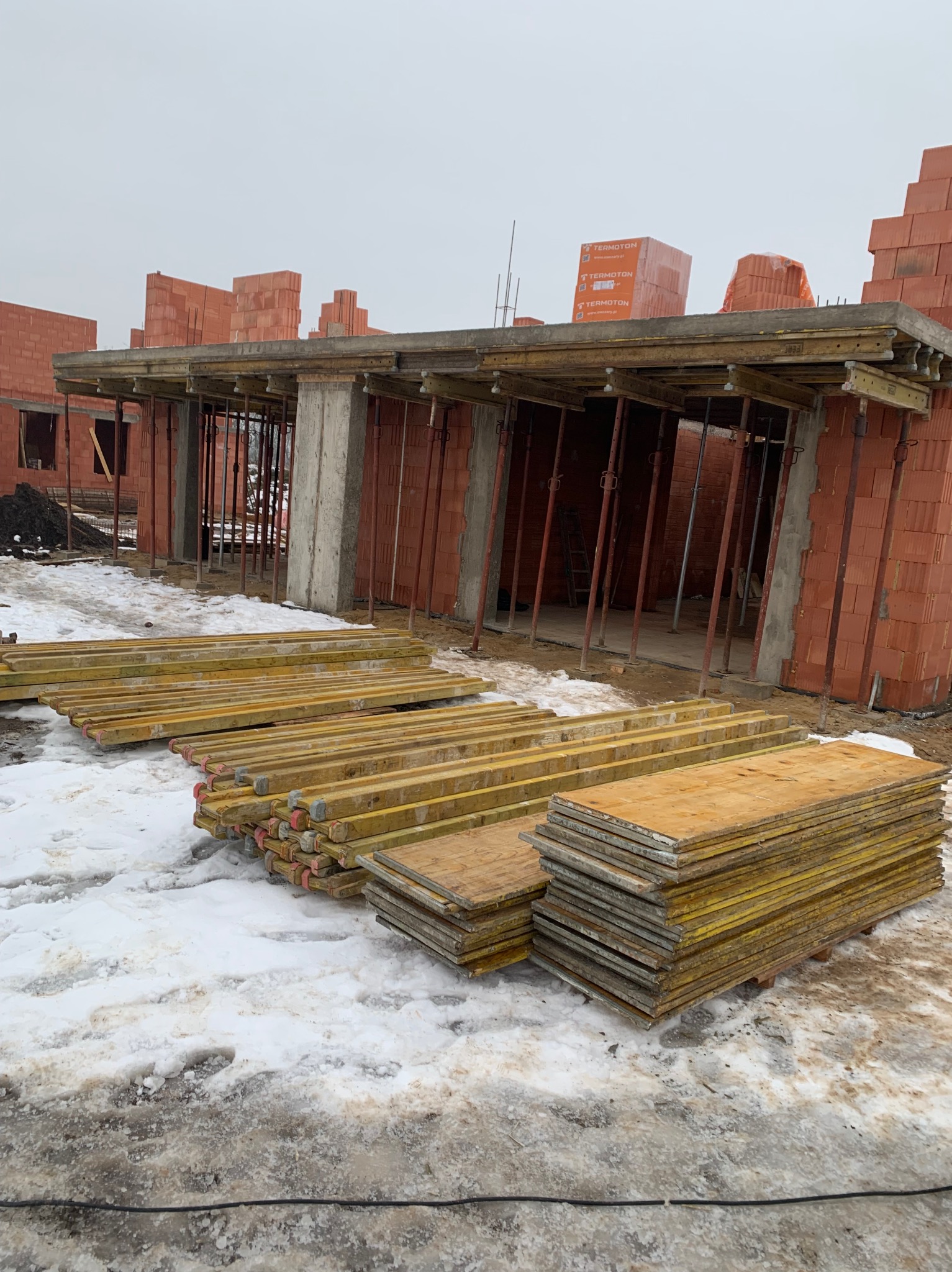 Wznoszenie ścian z czerwonej cegły na budowie w zimowy dzień. Widoczne drewniane szalunki i podpory. Cegły marki Termoton na górze konstrukcji.