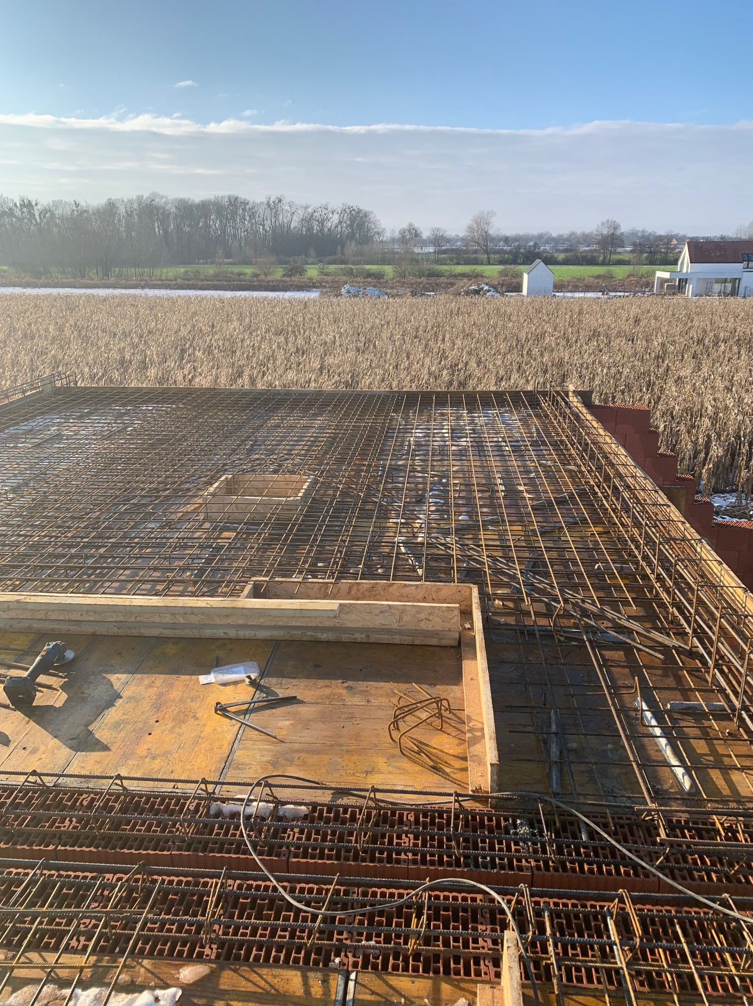 Zbrojenie płyty fundamentowej z widocznymi bloczkami i deskowaniem, na tle pola i zabudowań w oddali, w słoneczny dzień.