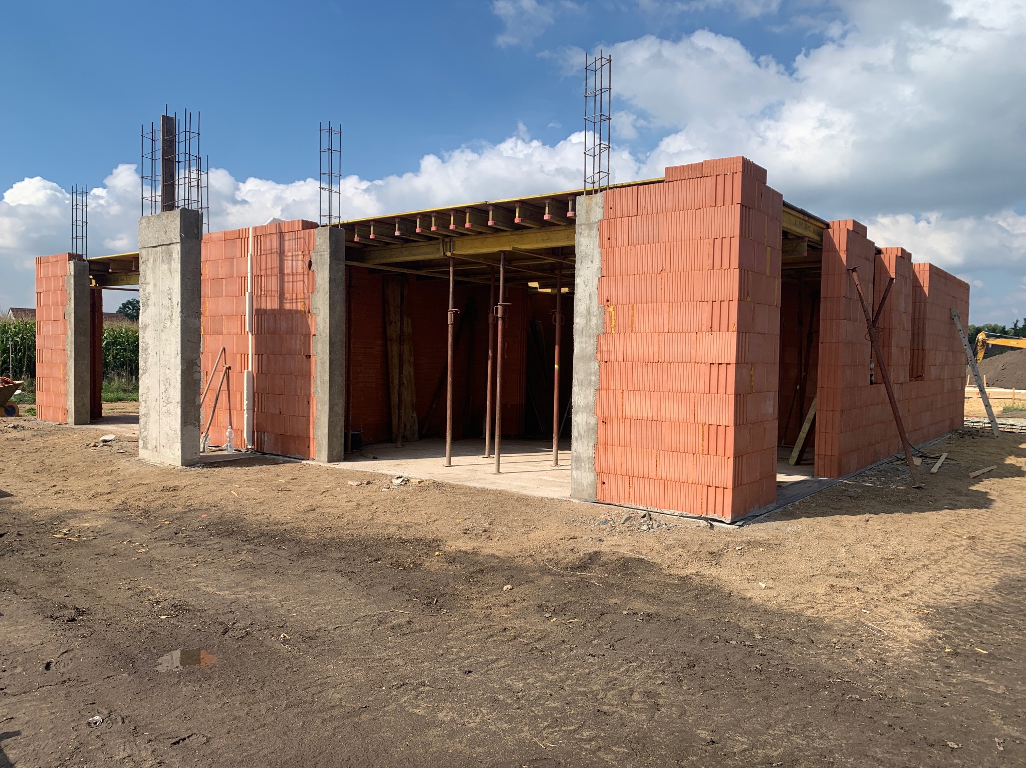 Wznoszenie ścian z czerwonej cegły na budowie domu, widoczne betonowe słupy i zbrojenia, drewniane szalunki stropu, ziemiste podłoże i błękitne niebo z chmurami w tle.