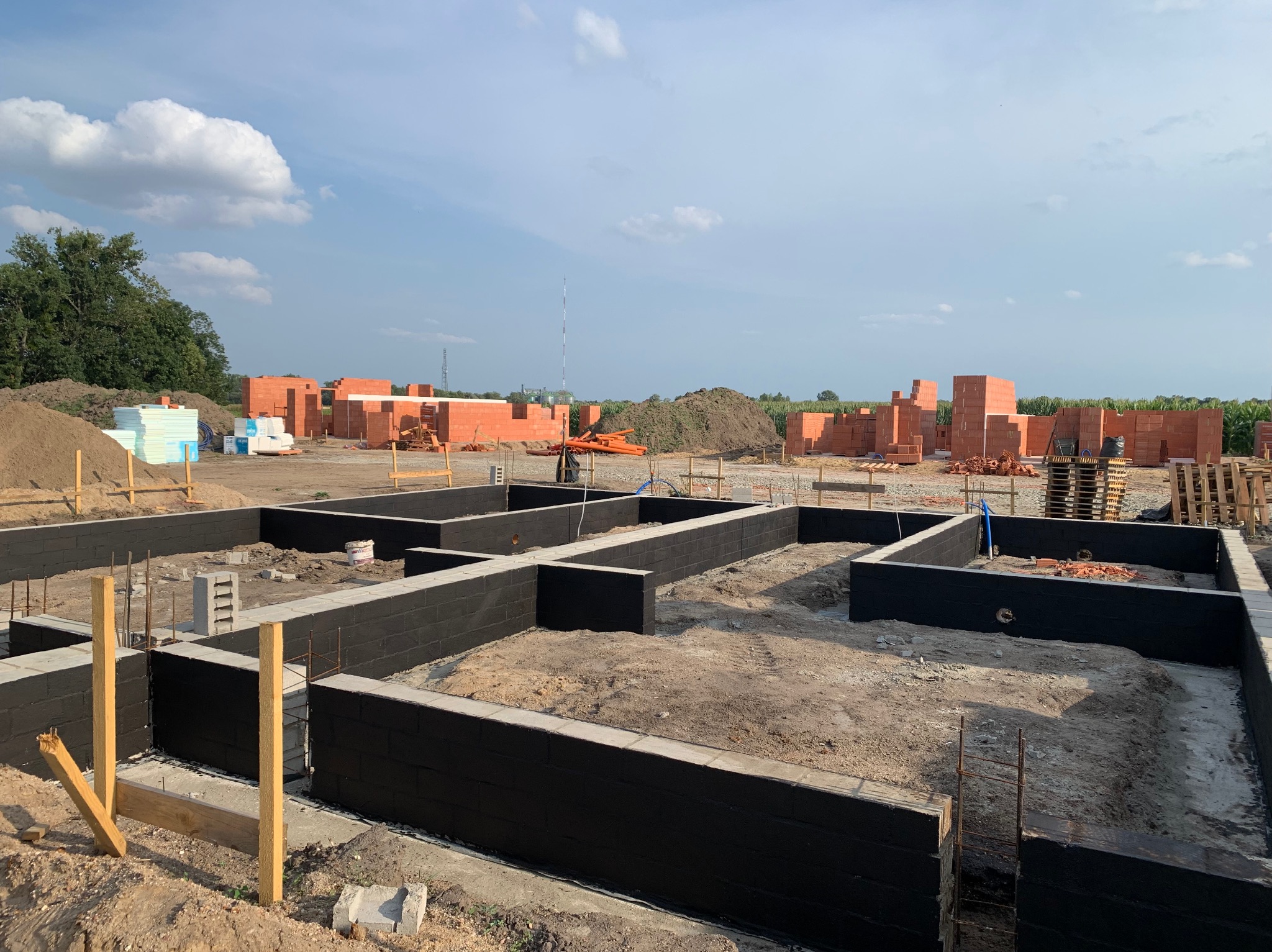 Zabezpieczone, czarną izolacją fundamenty domu jednorodzinnego, z widocznymi bloczkami z czerwonej cegły na dalszym planie i błękitnym niebem w tle.