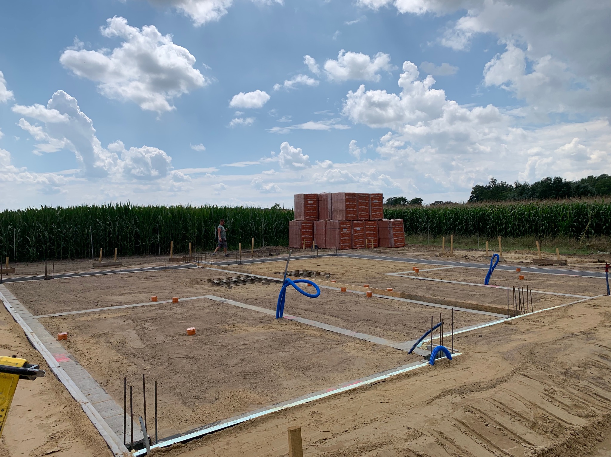 Przygotowany teren pod fundamenty z zaznaczonym obrysem, wystającymi prętami zbrojeniowymi i stosem cegieł w tle, otoczony polem kukurydzy pod błękitnym niebem.