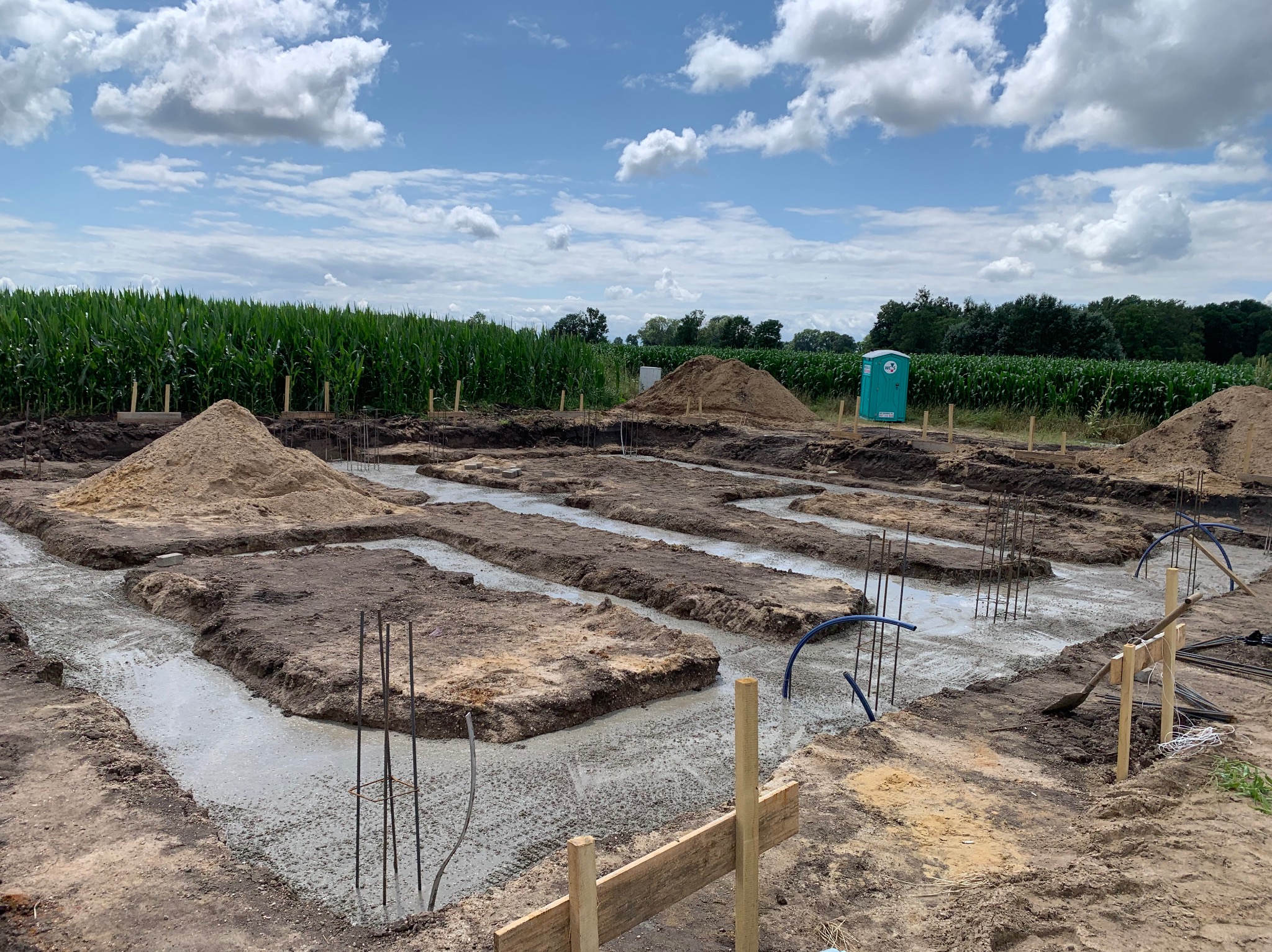 Wylane fundamenty pod budowę domu na tle pola kukurydzy i błękitnego nieba z chmurami. Widoczne zbrojenia, szalunki i przenośna toaleta.