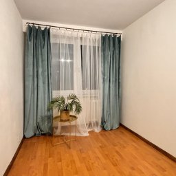 Monika Rakowska Usługi - Pusty pokój po home stagingu: drewniana podłoga, okno z zasłonami (turkusowe i białe), stolik z rośliną. Jasne ściany, minimalistyczny wystrój.