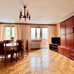 Monika Rakowska Usługi - Jasne wnętrze pokoju z parkietem, drewnianymi meblami i stołem z kwiatami. Widok na okno z zasłonami i balkon. Ciepłe oświetlenie.