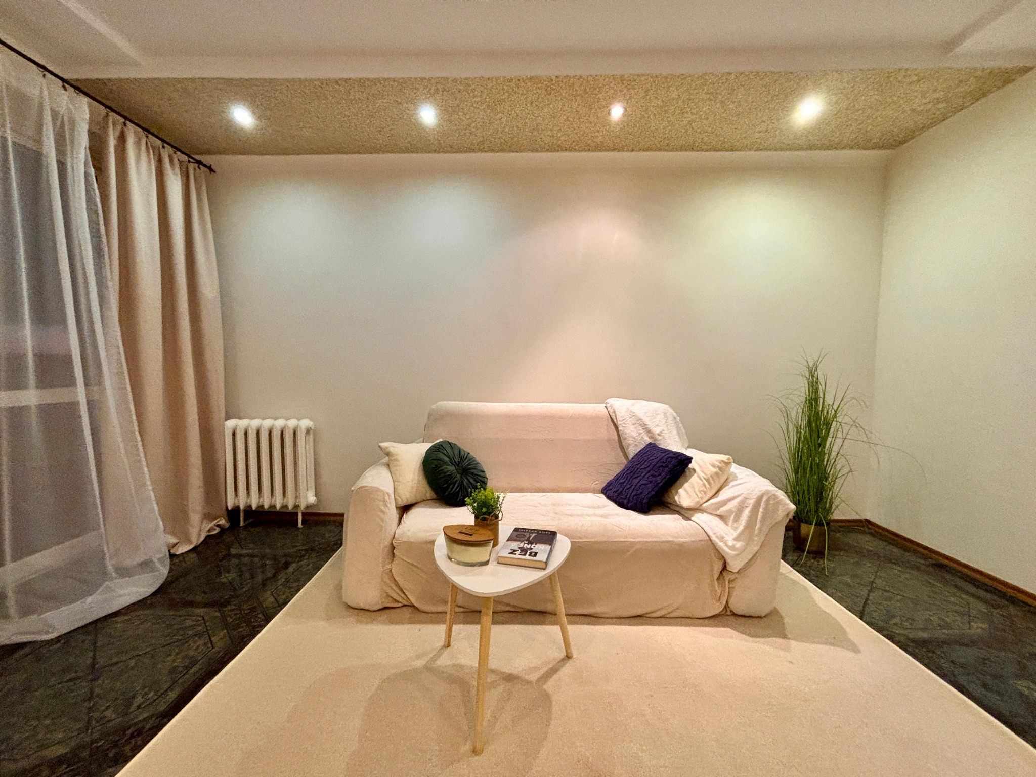 Jasny salon po home stagingu: beżowa sofa z poduszkami, stolik z książką, dywan w odcieniach beżu, zasłony i grzejnik. Wnętrze z rośliną doniczkową.