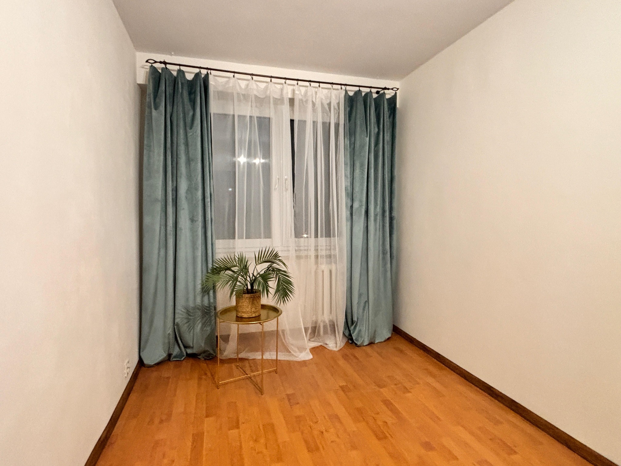 Pusty pokój po home stagingu: drewniana podłoga, okno z zasłonami (turkusowe i białe), stolik z rośliną. Jasne ściany, minimalistyczny wystrój.