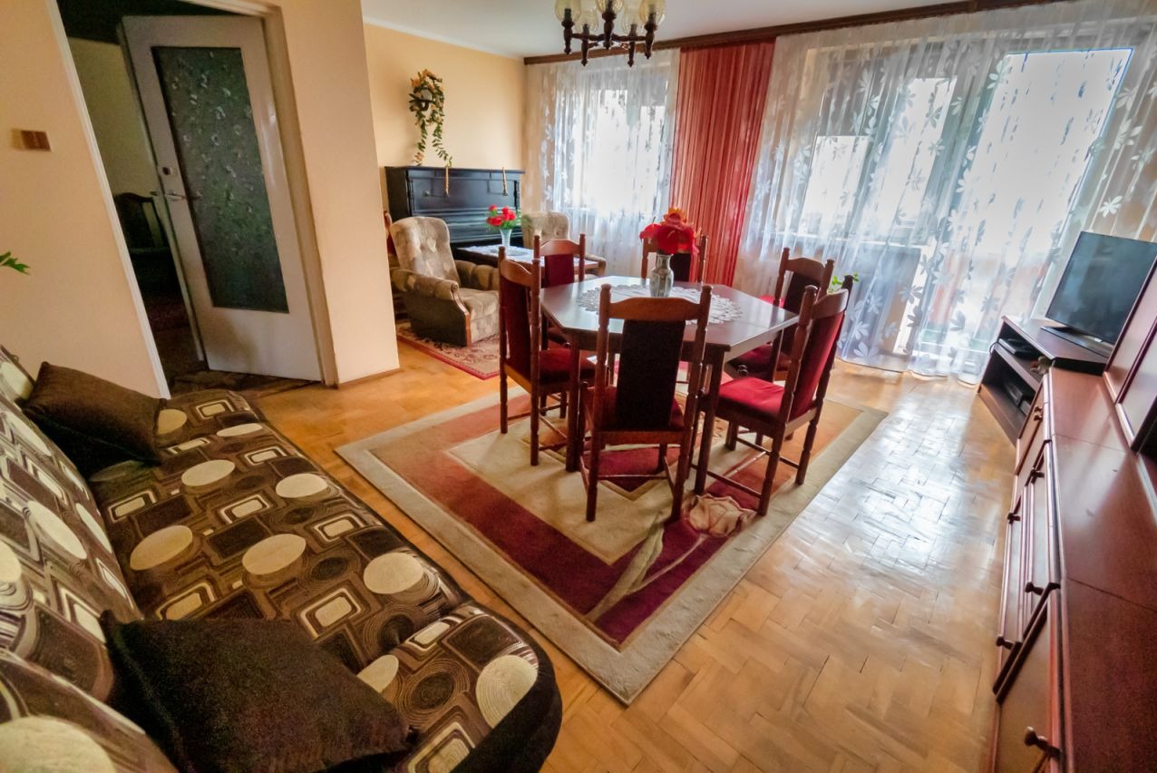 Salon z pianinem, stołem i krzesłami w stylu vintage, dywanem z motywem kwiatowym i parkietem. Widok na okno z firankami i telewizor na komodzie.