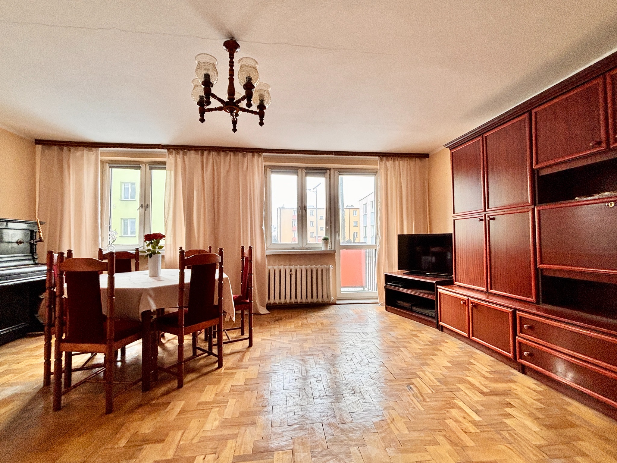 Jasne wnętrze pokoju z parkietem, drewnianymi meblami i stołem z kwiatami. Widok na okno z zasłonami i balkon. Ciepłe oświetlenie.