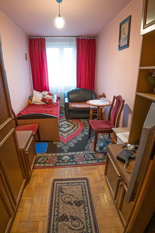 Mały pokój z łóżkiem, sofą i dywanem w stylu vintage. Widok na wnętrze przed home stagingiem, z meblami w stylu retro i oświetleniem punktowym.