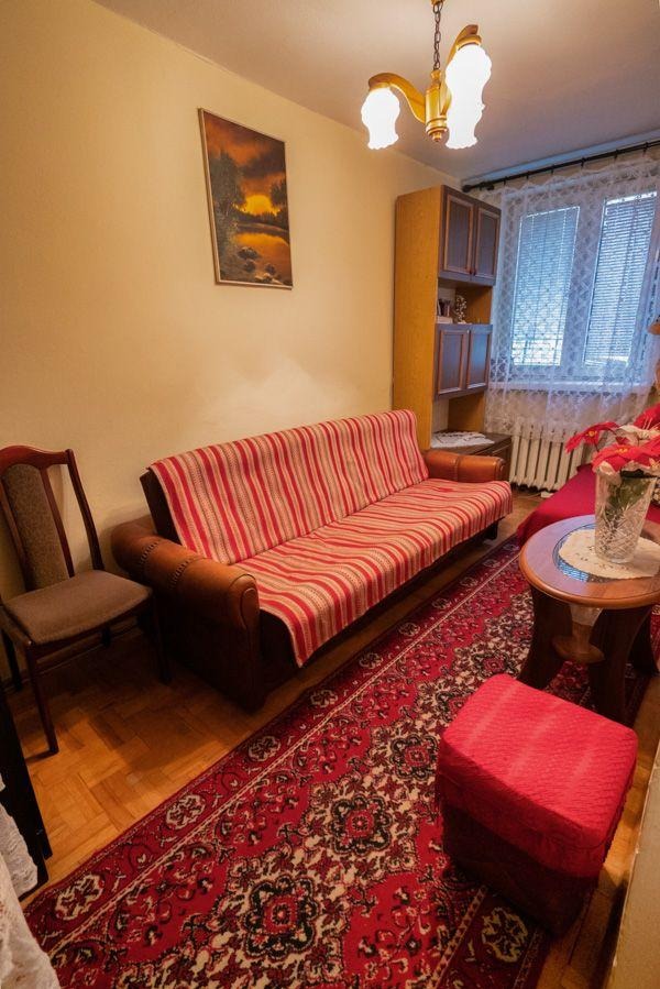 Pokój dzienny przed home staging: sofa w paski, czerwony dywan z wzorami, drewniana podłoga i meble. Widok na okno z firanką i obraz na ścianie.