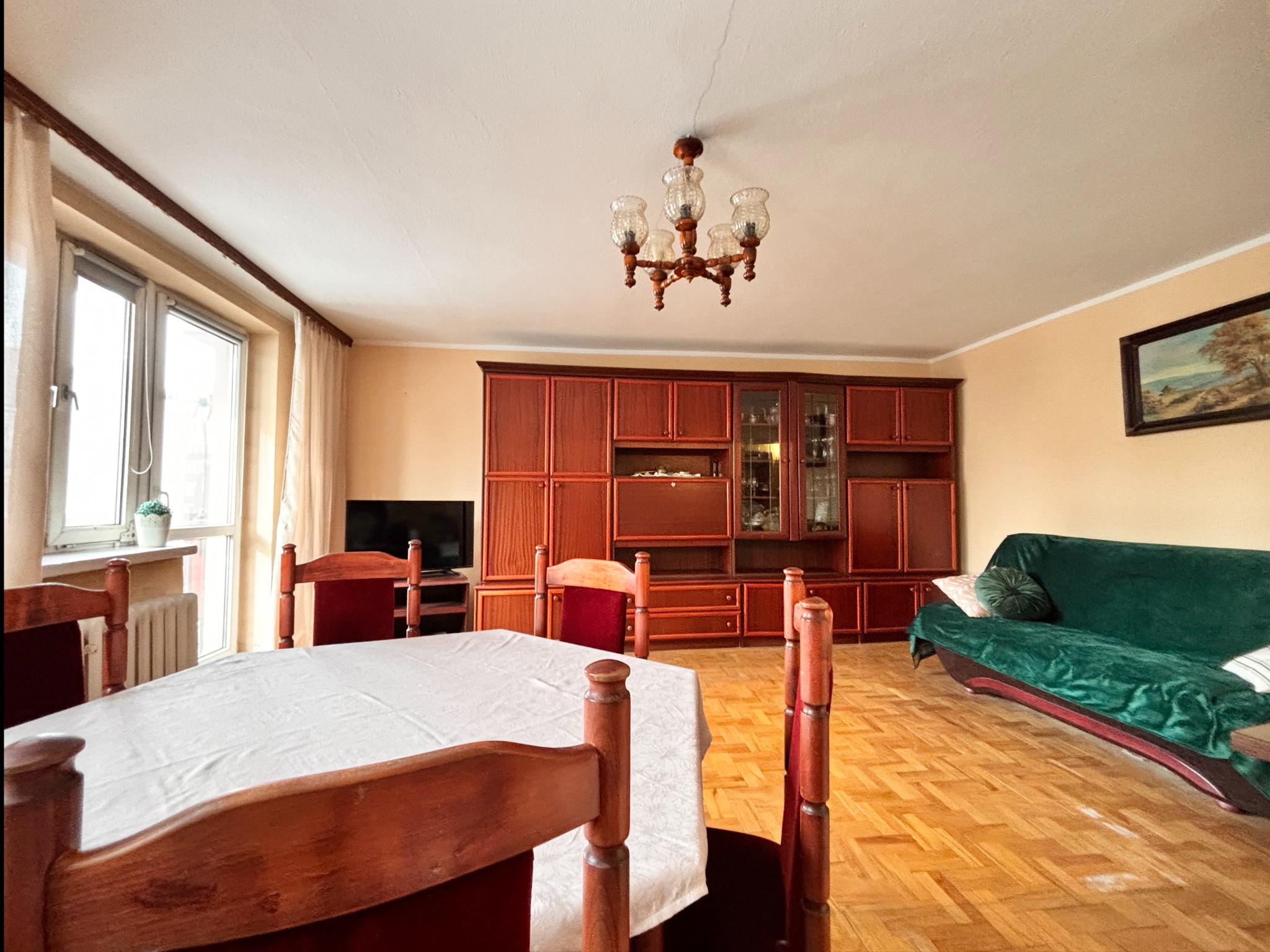 Jasne wnętrze po home staging: stół z białym obrusem, drewniane krzesła, parkiet, zielona sofa i duża, ciemna meblościanka. Widok na okno z kwiatem.