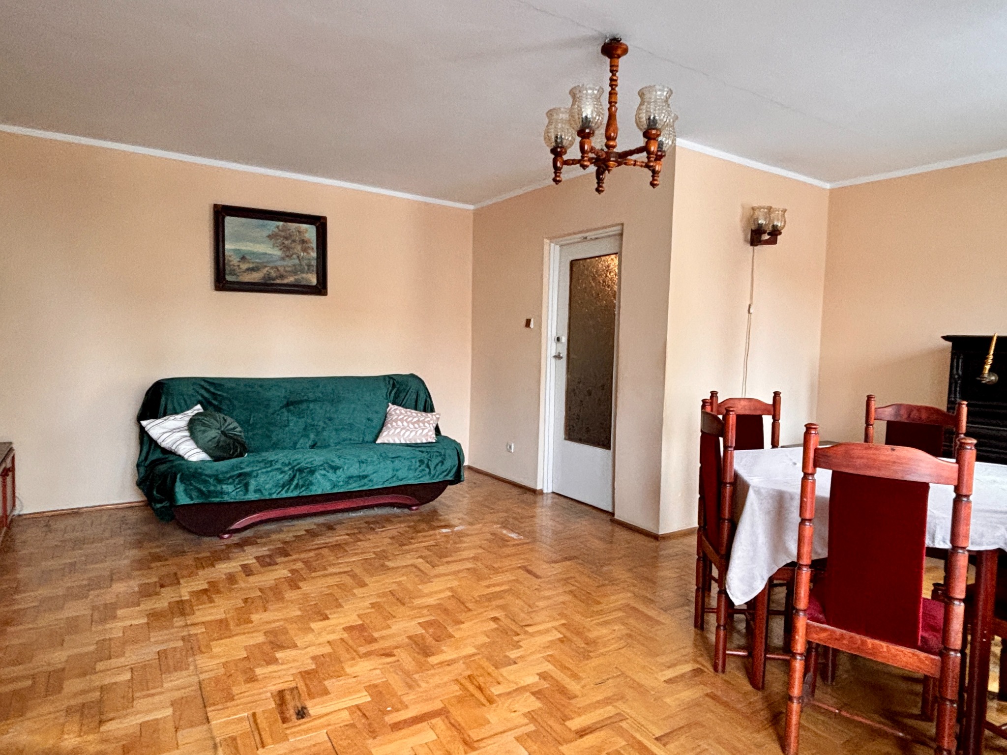 Salon po home staging: parkiet w jodełkę, zielona sofa z poduszkami, drewniany stół z krzesłami, lampa wisząca i obraz na ścianie w ciepłych barwach.