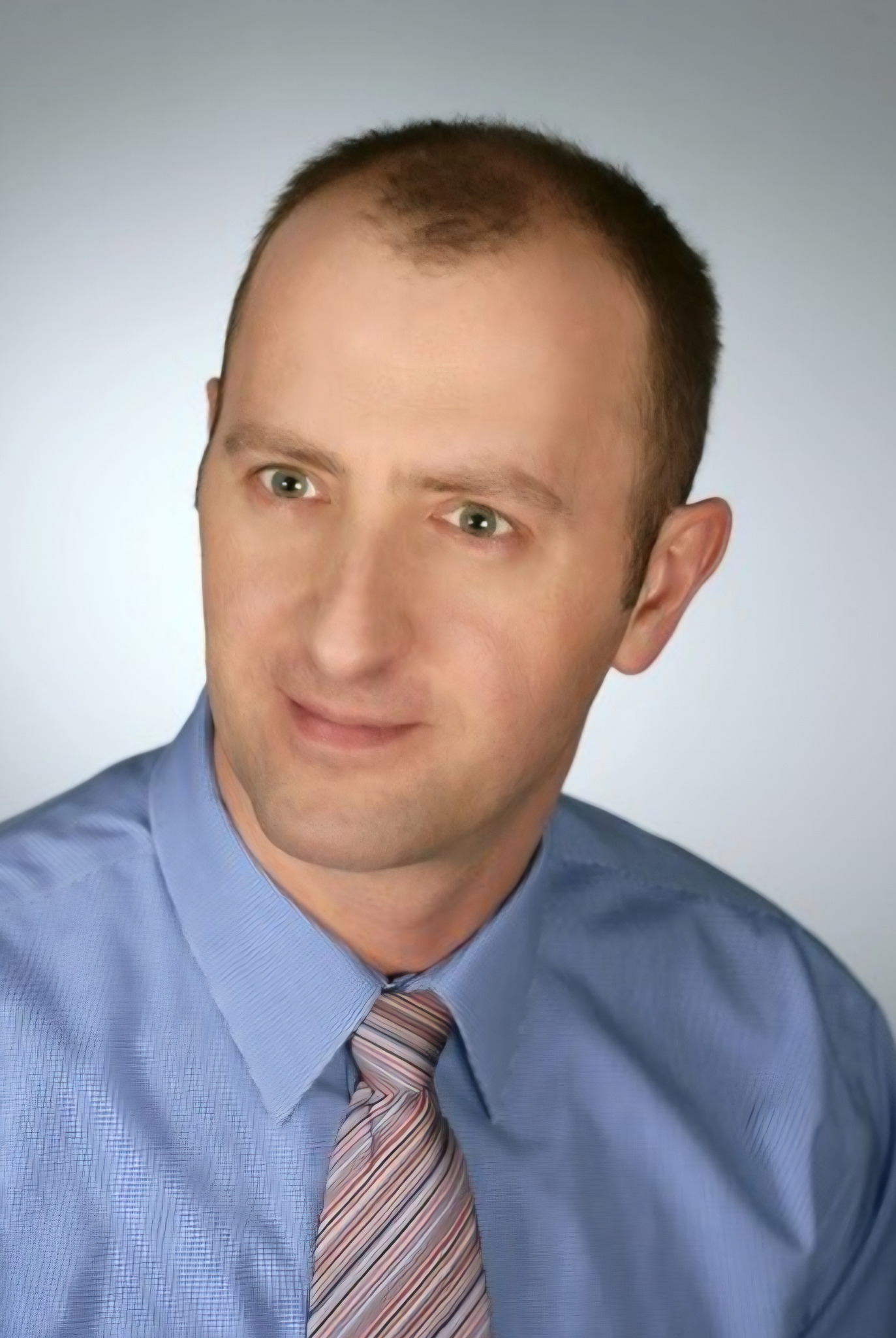 Portret mężczyzny w błękitnej koszuli i krawacie w paski, z delikatnym uśmiechem i spojrzeniem w bok, na jasnym tle. Zdjęcie biznesowe.