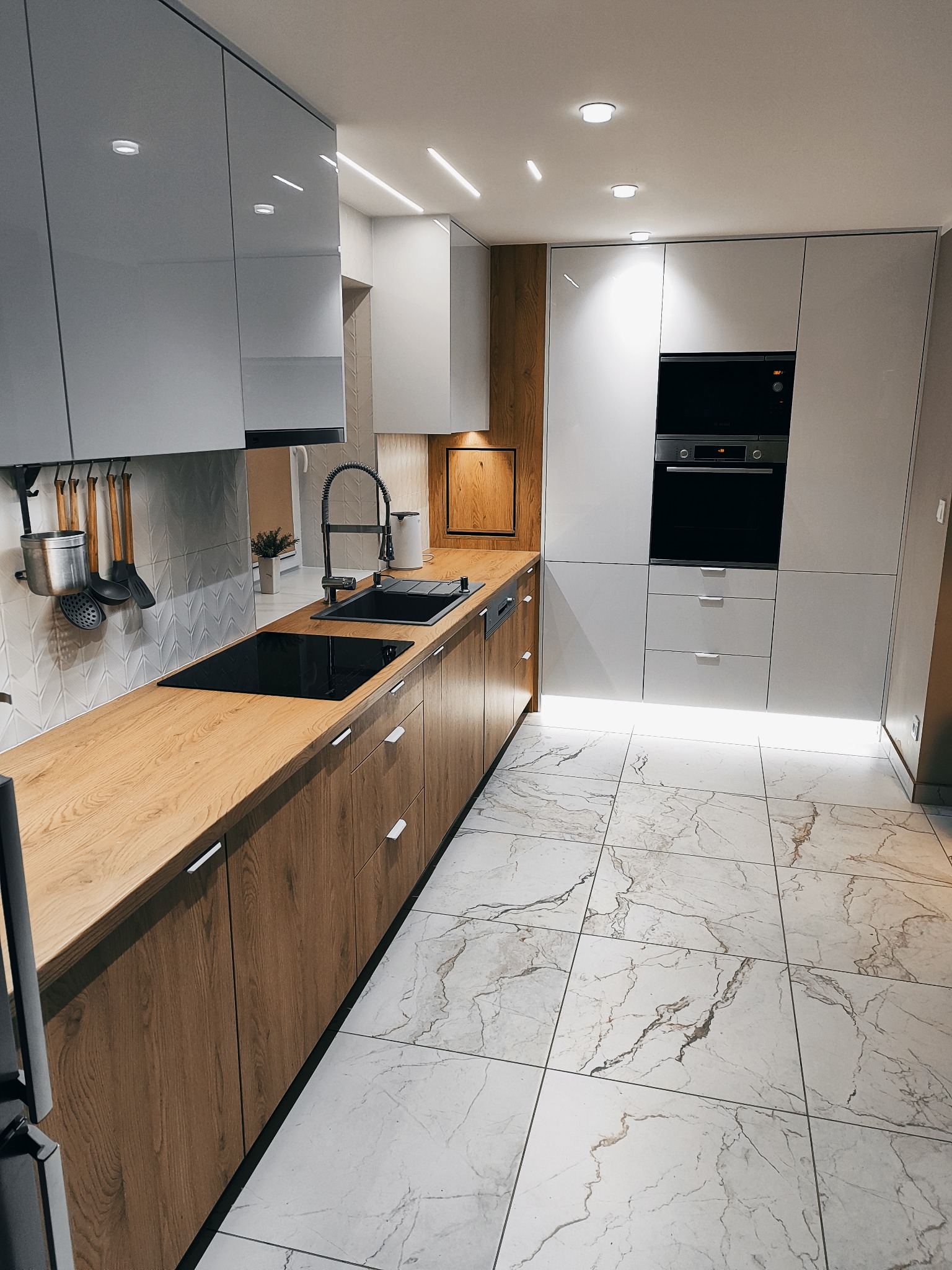 Nowoczesna kuchnia z drewnianymi szafkami dolnymi i białymi górnymi, marmurowa podłoga, czarny zlew i płyta indukcyjna. Oświetlenie LED pod szafkami.