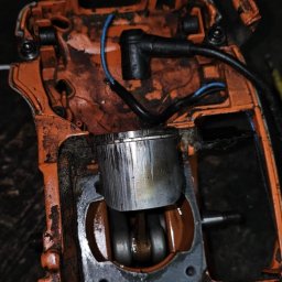 Liwiusz Stoppa - Pomarańczowa obudowa piły spalinowej z widocznym cylindrem i tłokiem, detale zużycia i zabrudzenia, zbliżenie na mechanizm w trakcie naprawy.