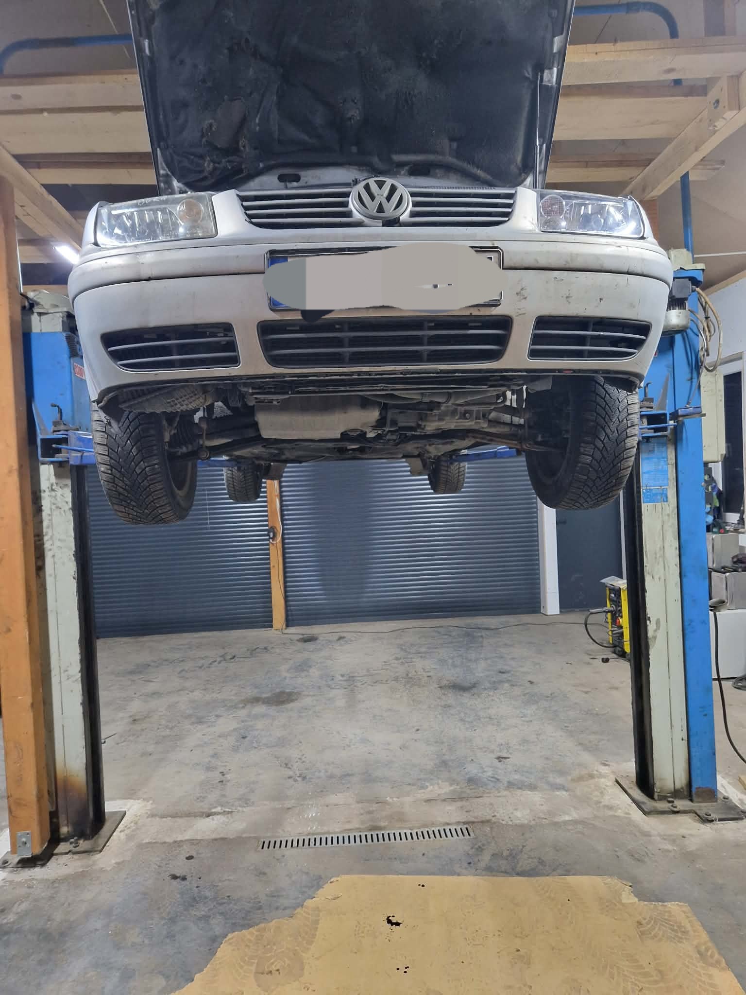 Podnośnik samochodowy z uniesionym srebrnym Volkswagenem Golfem z otwartą maską w warsztacie. Widoczne opony zimowe i podwozie pojazdu.