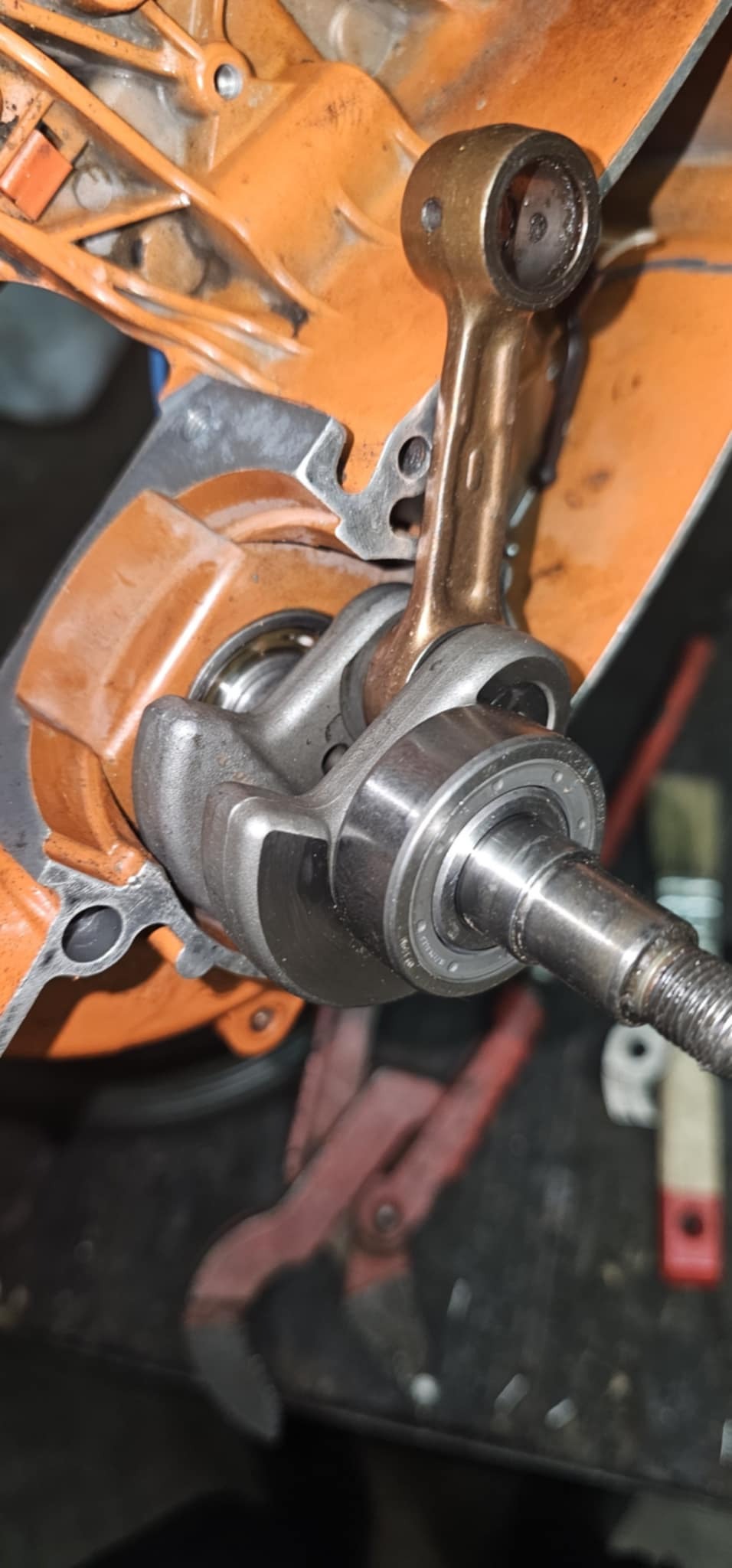 Rozłożony silnik spalinowy z widocznym wałem korbowym, tłokiem i łożyskami. Pomarańczowa obudowa silnika, narzędzia w tle. Precyzyjna naprawa silnika.