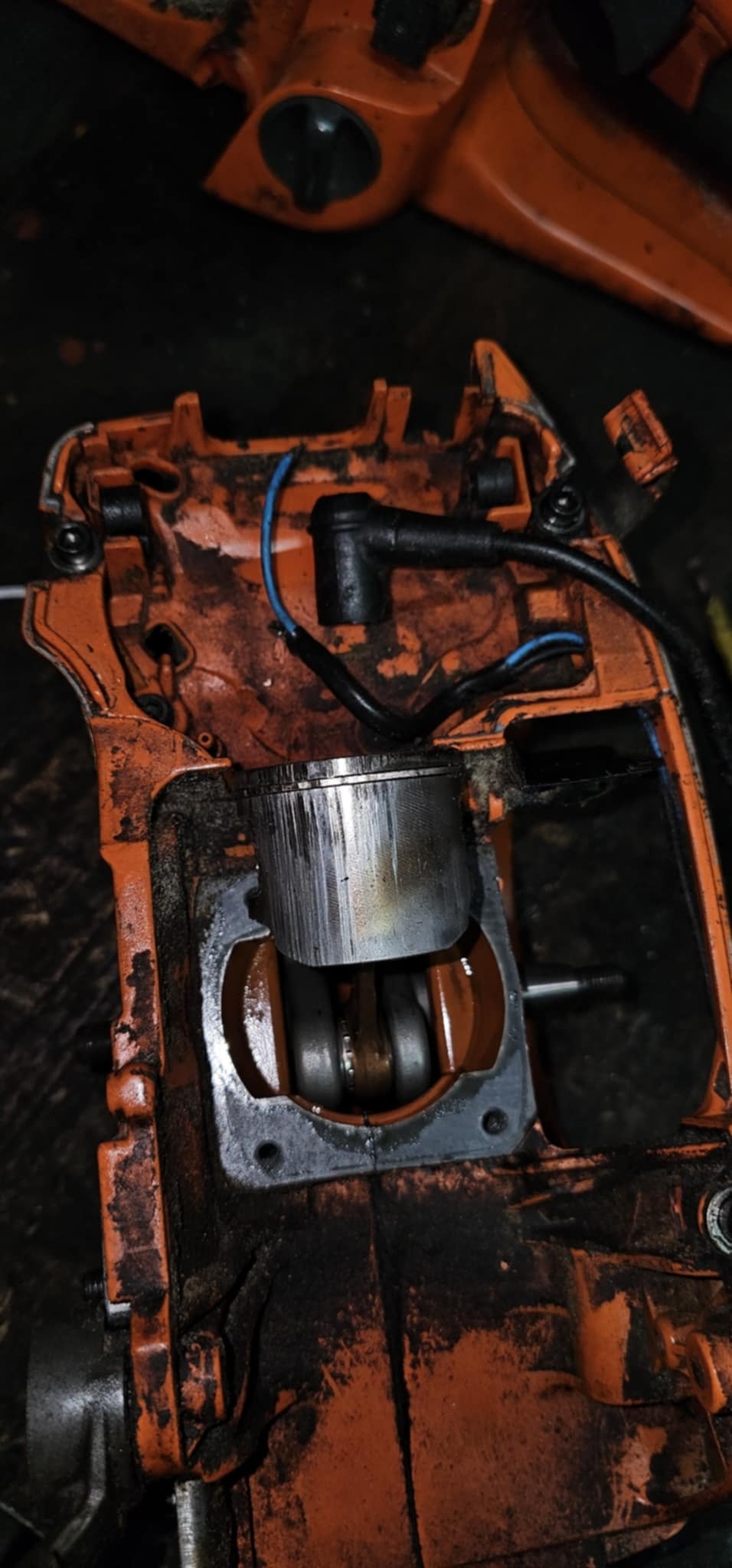 Pomarańczowa obudowa piły spalinowej z widocznym cylindrem i tłokiem, detale zużycia i zabrudzenia, zbliżenie na mechanizm w trakcie naprawy.