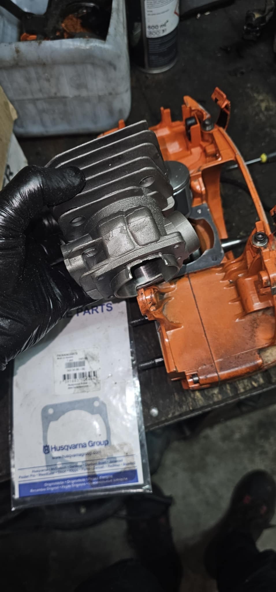 Cylinder silnika spalinowego w dłoni w czarnej rękawicy, obok korpus pomarańczowej pilarki Husqvarna i opakowanie z uszczelkami. Widoczne ślady użytkowania.