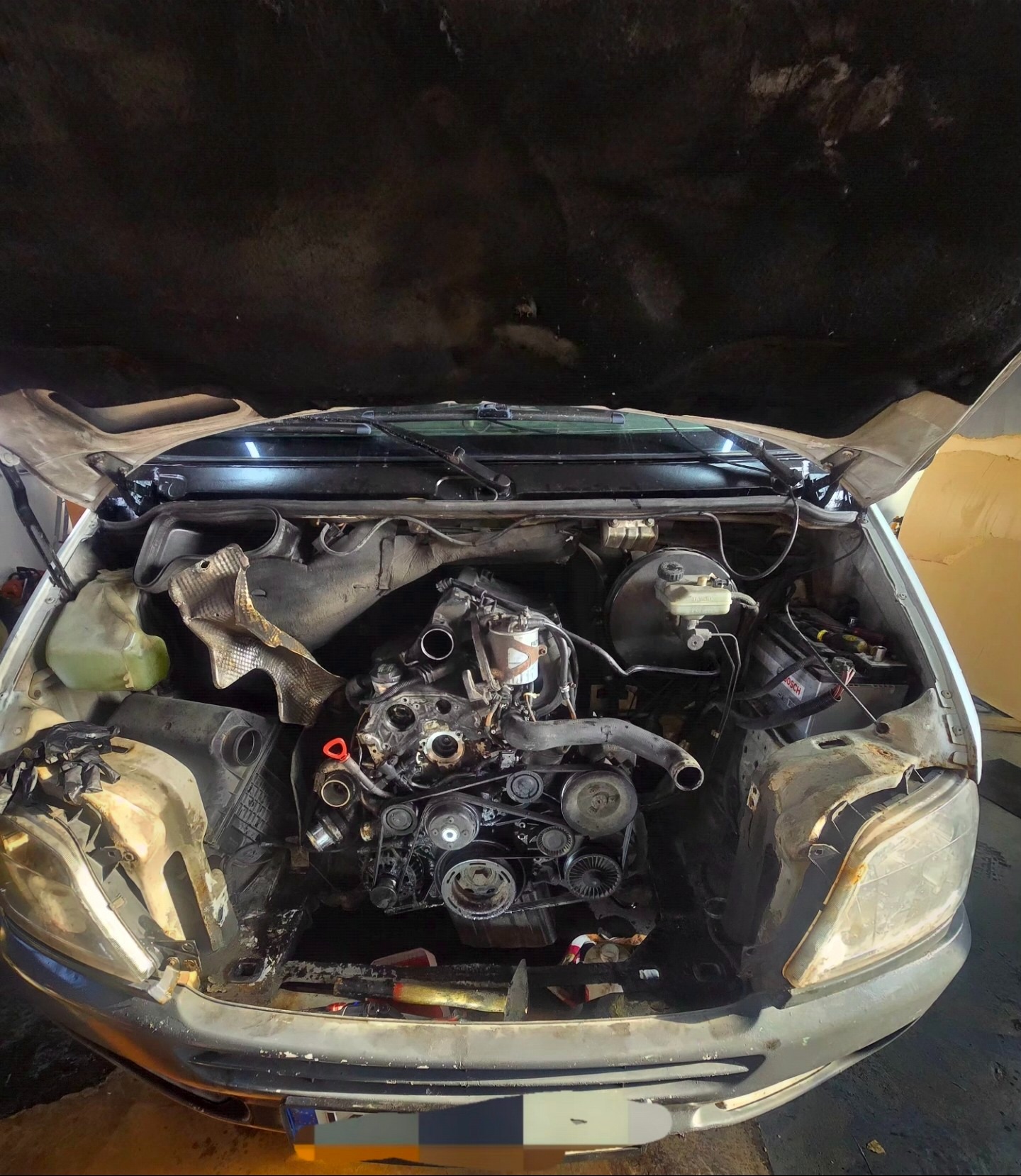 Widok z przodu na silnik samochodu w trakcie naprawy, z otwartą maską, ukazujący skomplikowaną mechanikę i narzędzia w warsztacie. Detaliczna prezentacja procesu serwisowego.